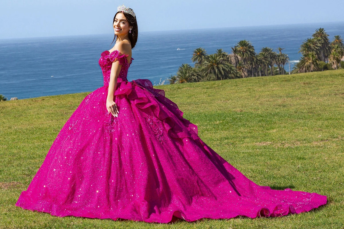 Shown in a angled view, the Fuchsia quinceañera dress has off-shoulder neckline and a tiered glitter skirt with a train.