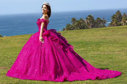 Shown in a angled view, the Fuchsia quinceañera dress has off-shoulder neckline and a tiered glitter skirt with a train.