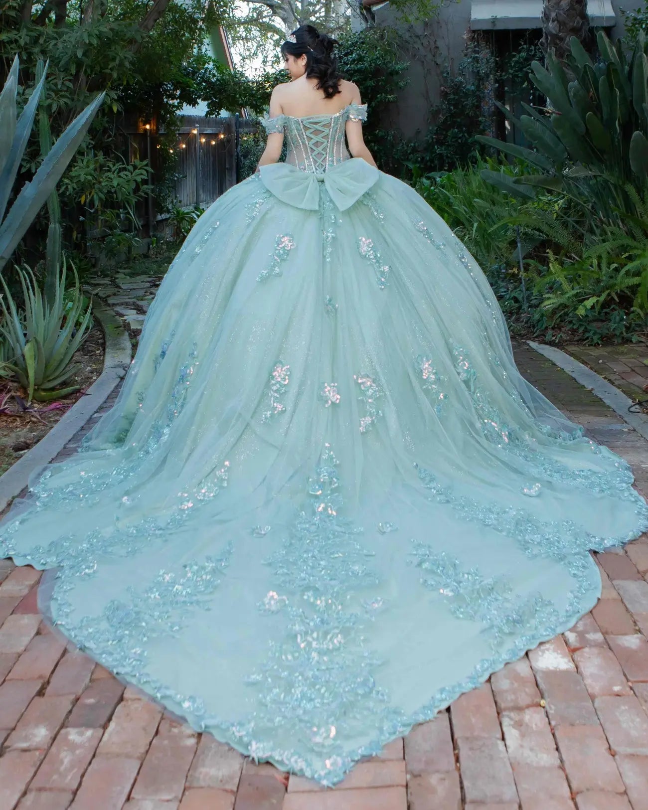 Back view of a silver sage quinceanera dress with lace-up corset, voluminous tulle bow, 3D floral appliques, and chapel-length train