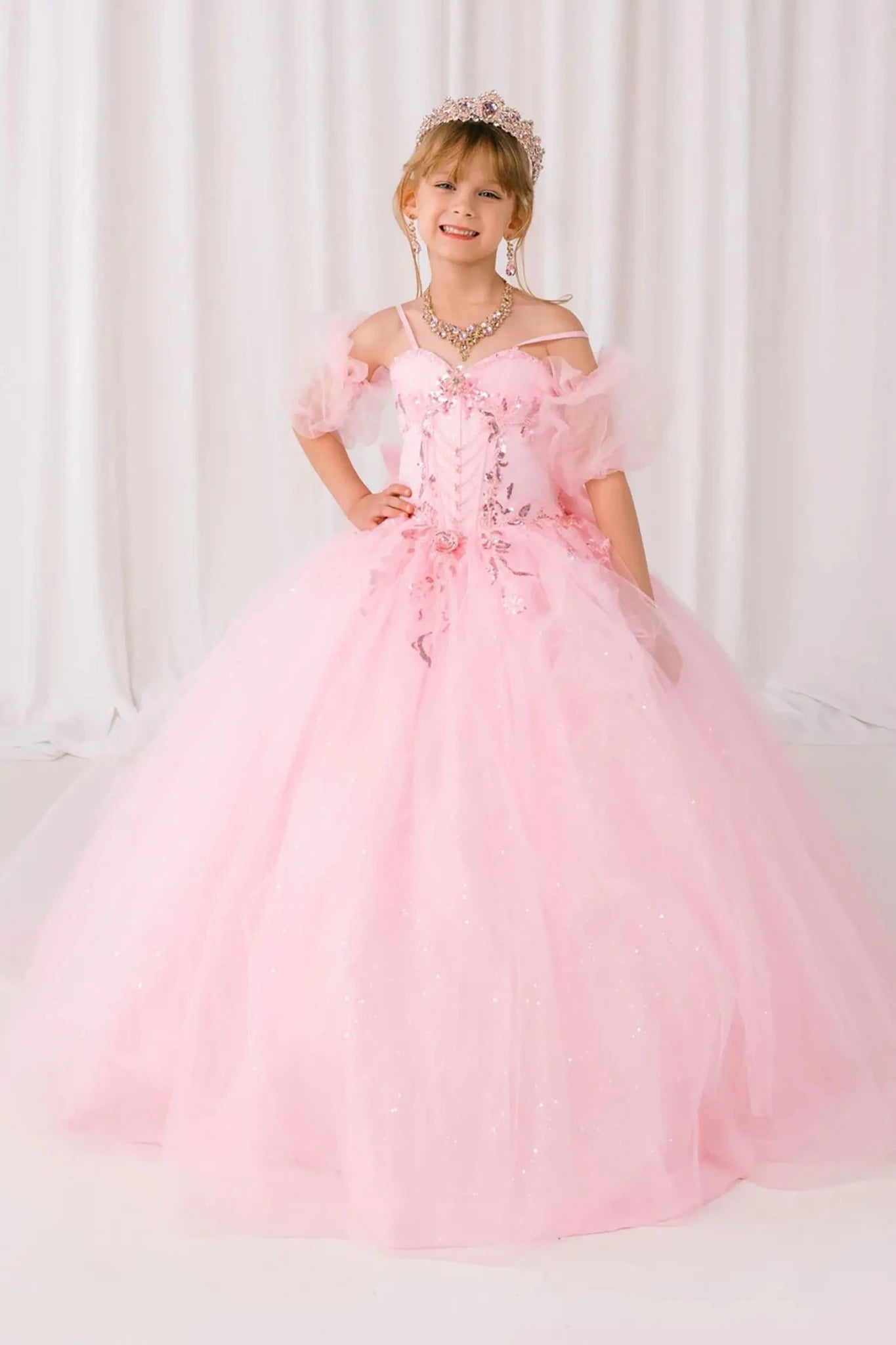 Young girl standing front-facing in burgundy mini quinceañera ball gown with beaded bodice, floral appliqué, draped details, sparkling tulle skirt, and matching tulle arm cuffs