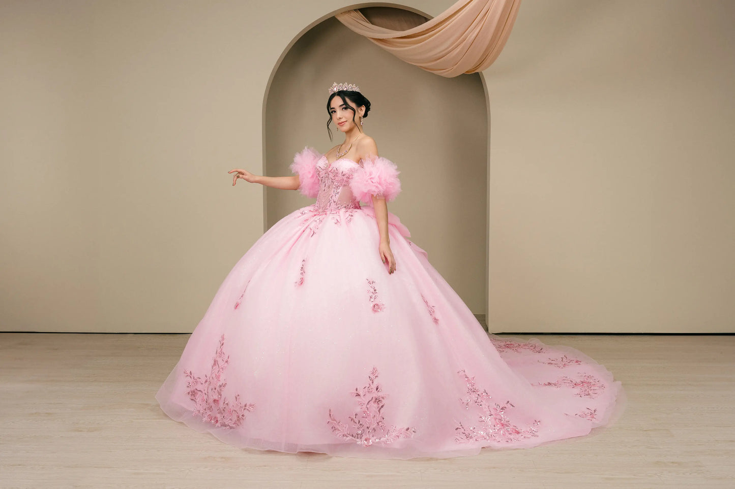 Angled front view:
Pink off-shoulder quinceañera ball gown featuring floral appliqué details, voluminous skirt, and fitted corset bodice in a three-quarter front view.