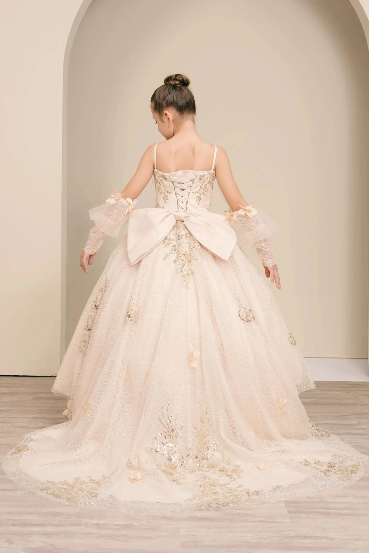 Back full-length view of Champagne mini quince mini quince dress with glitter floral embroidered bodice, full skirt, lace up back, oversized bow, and sheer lace gloves.