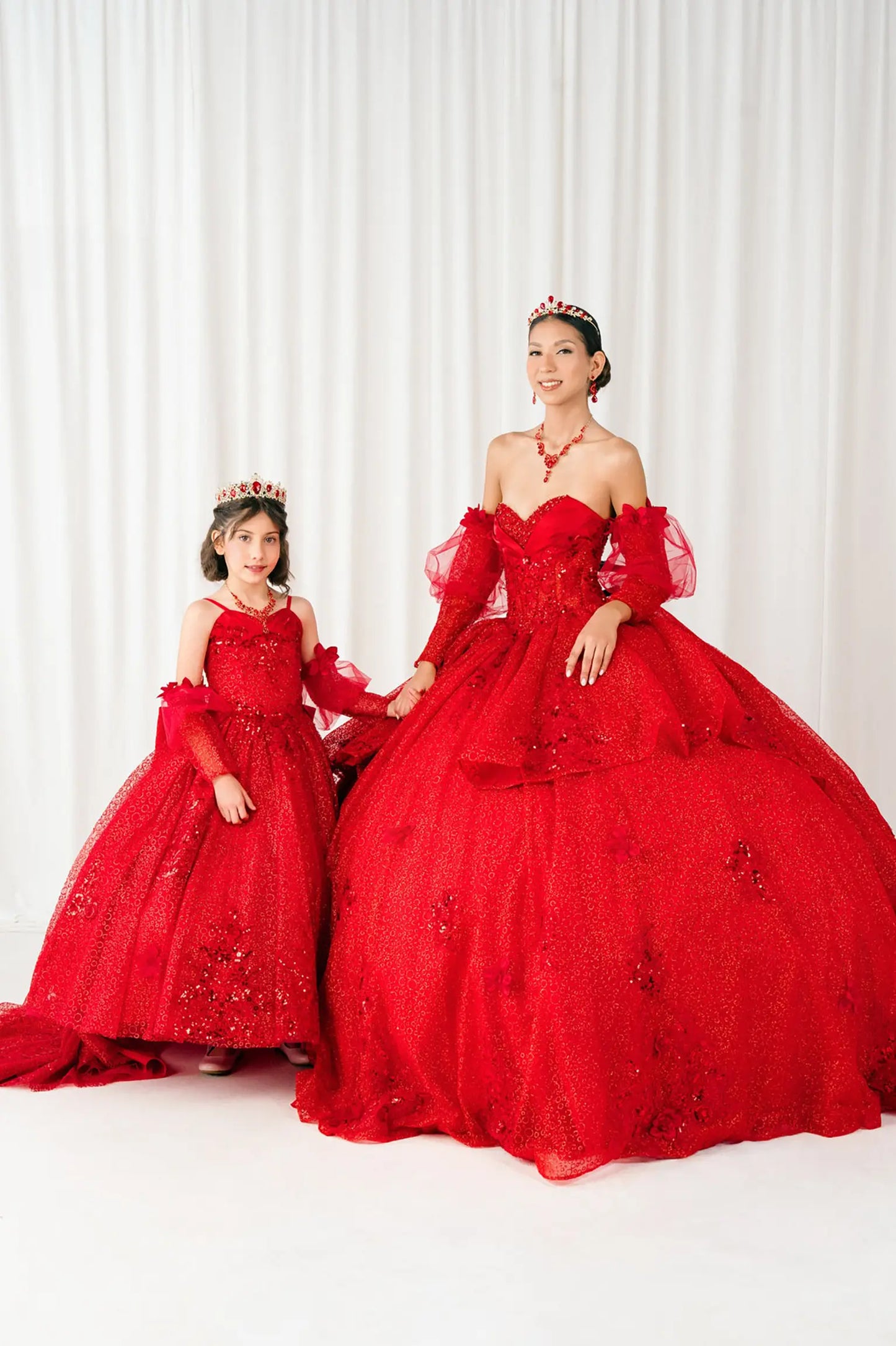 Full front view of a woman and kid wearing red quinceañera dress with glitter tulle skirt, floral applique bodice, and detachable sheer tulle sleeves.