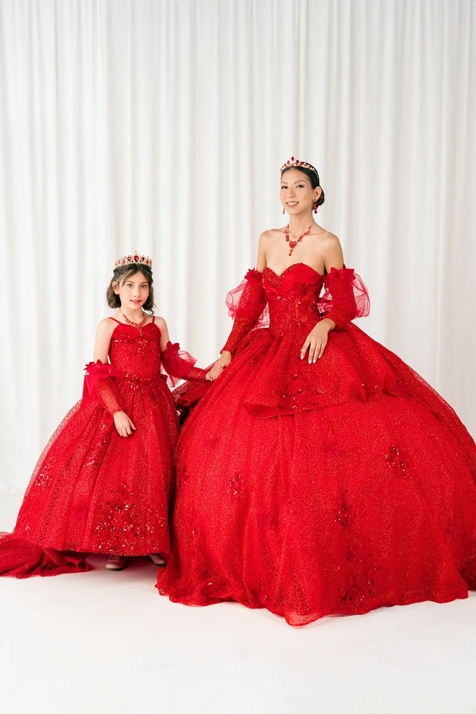 Full front view of a woman and kid wearing red quinceañera dress with glitter tulle skirt, floral applique bodice, and detachable sheer tulle sleeves.