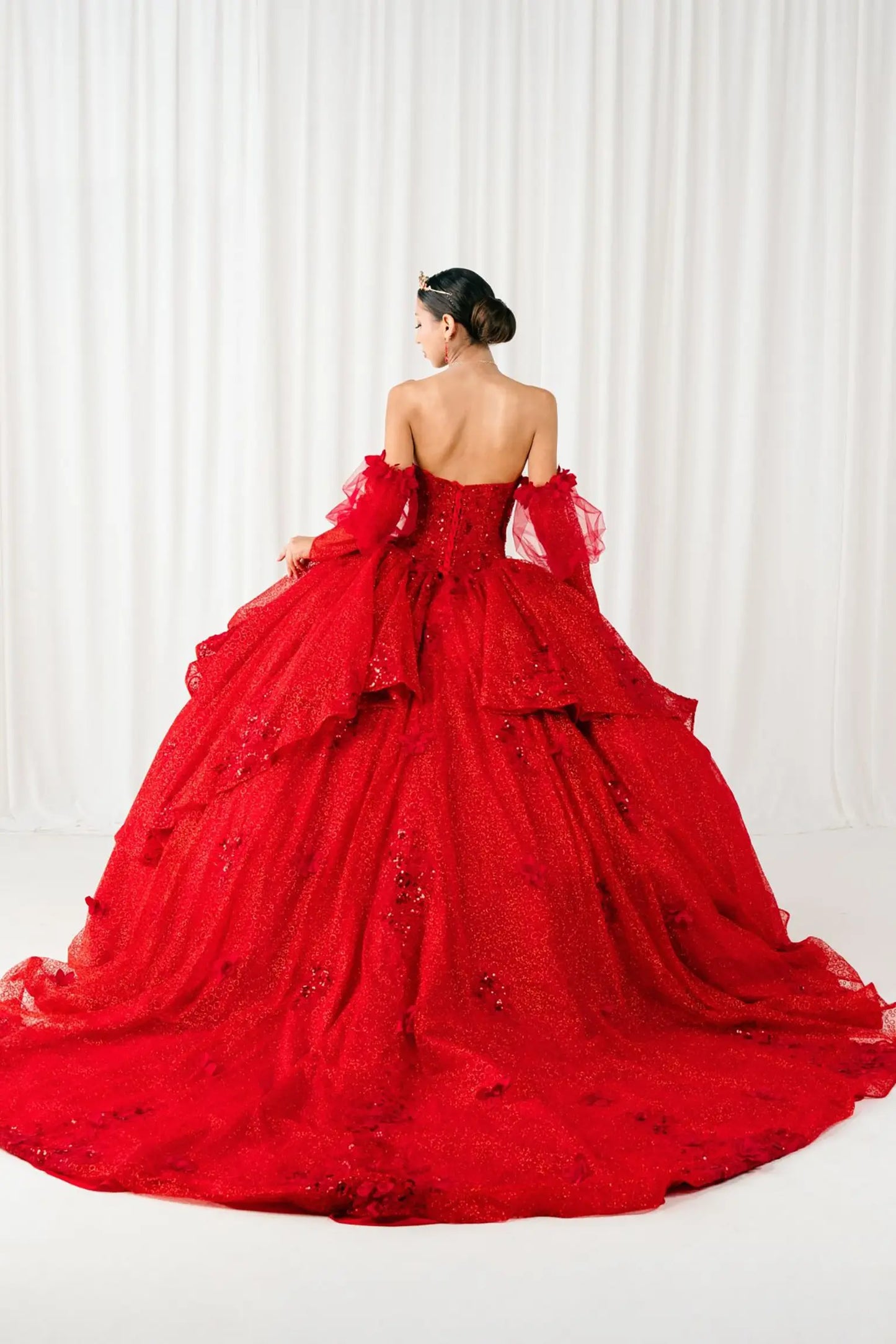 Full back view of a woman wearing red quinceañera dress with voluminous glitter tulle skirt, lace-up corset closure, and floral applique embellishments.