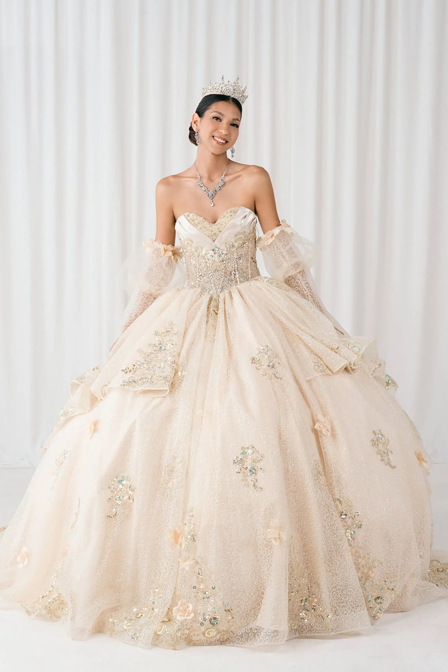 Front full-length view of Champagne quinceañera ball gown with off shoulder tulle sleeves, sweetheart sequin bodice, layered glitter tulle skirt, and cathedral train