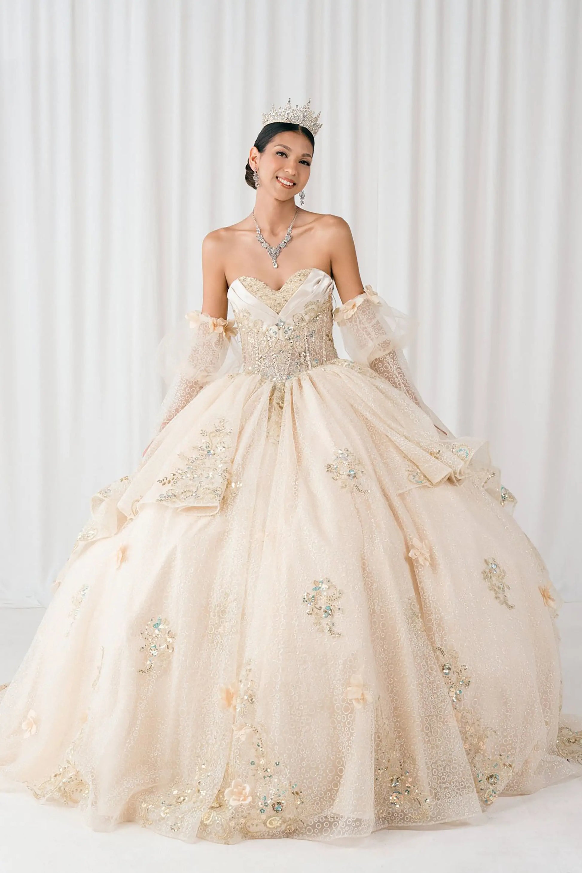 Front full-length view of Champagne quinceañera ball gown with off shoulder tulle sleeves, sweetheart sequin bodice, layered glitter tulle skirt, and cathedral train