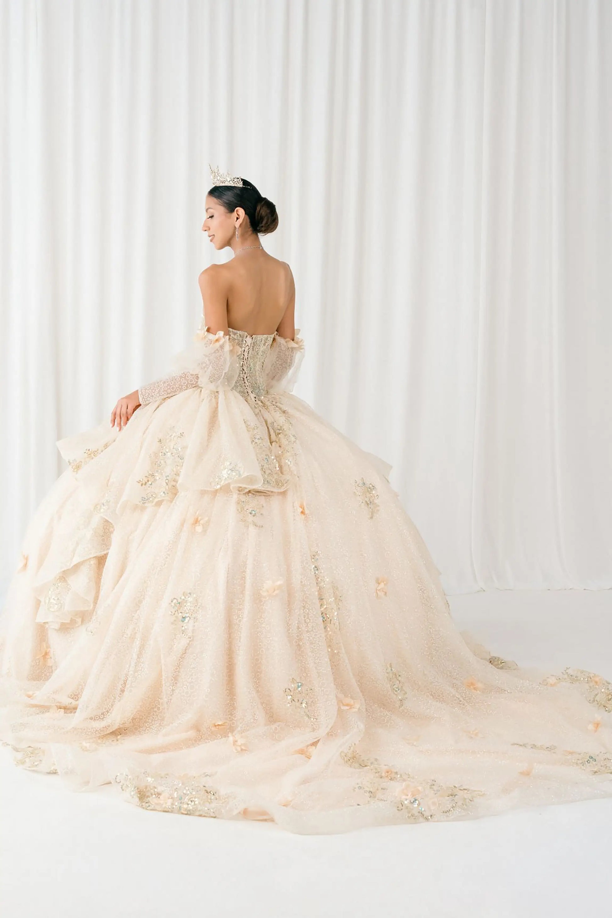 Side full-length view of Champagne quinceañera ball gown with off shoulder tulle sleeves, sweetheart sequin bodice, layered glitter tulle skirt, and cathedral train