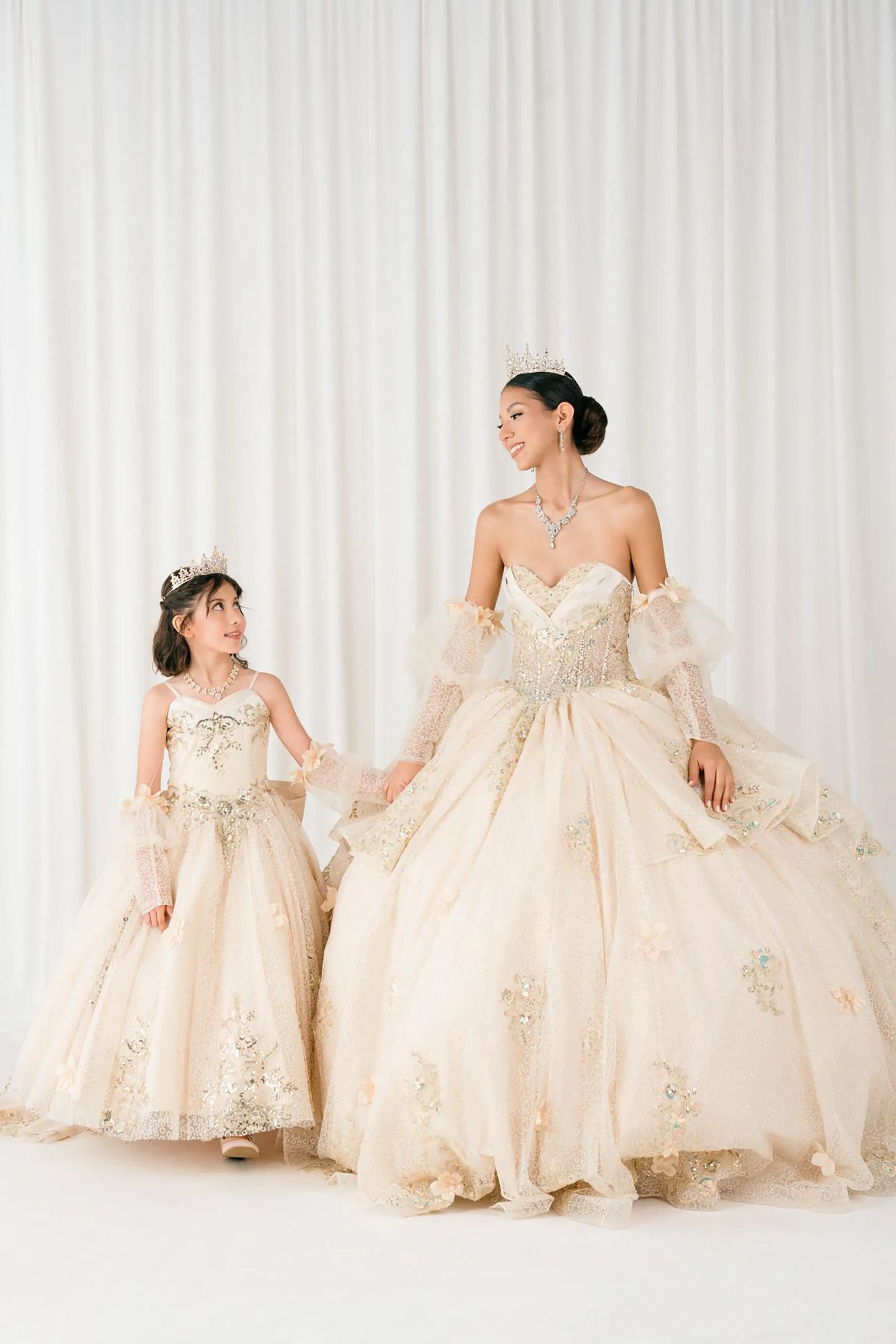 Champagne quinceañera and mini quinceañera ball gown with off shoulder tulle sleeves, sweetheart sequin bodice, layered glitter tulle skirt, and cathedral train