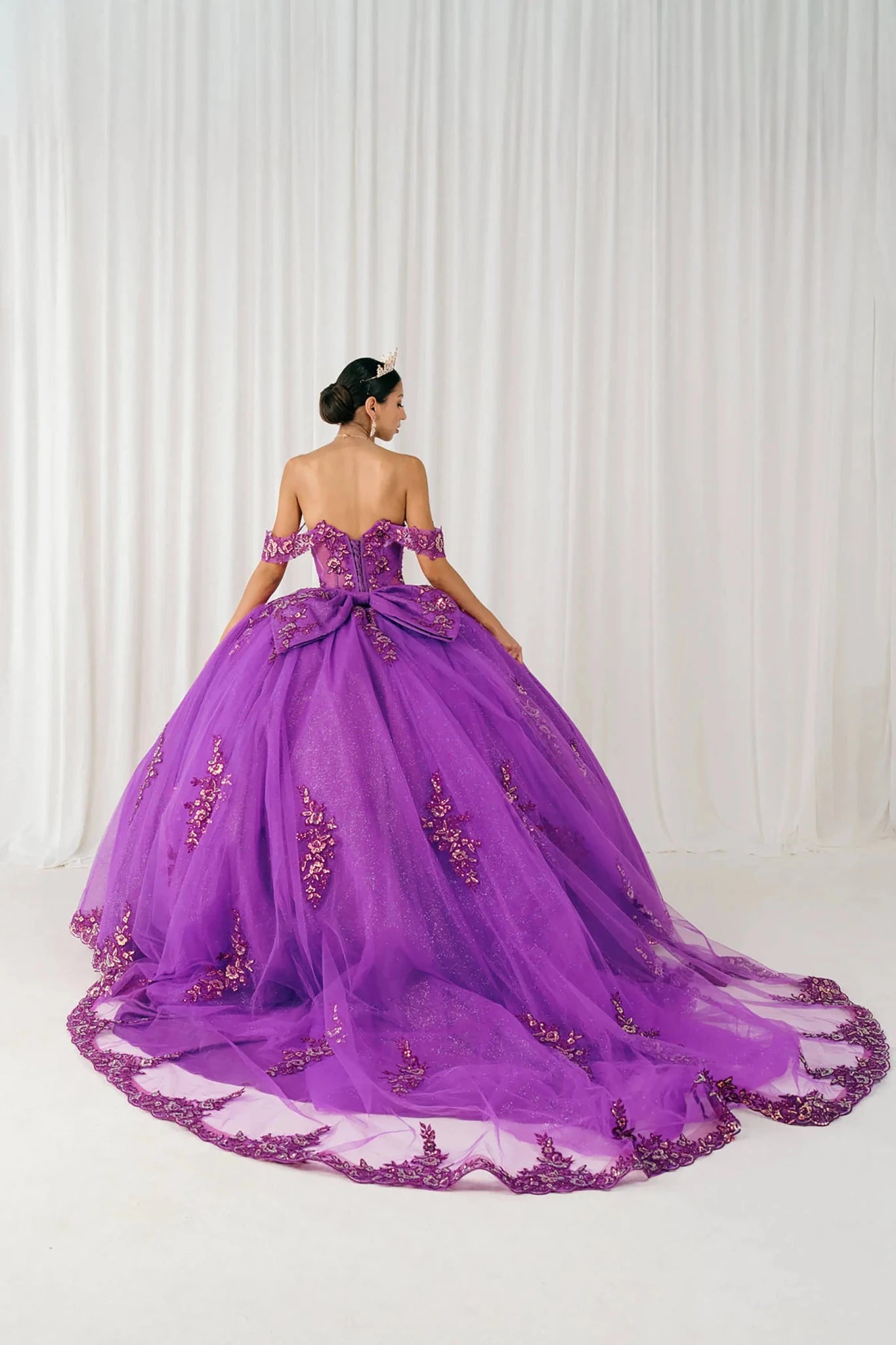 Back full-length view of Magenta Off shoulder quinceañera ball gown with beaded corset, floral appliqués, layered tulle skirt, cathedral length train, and bow