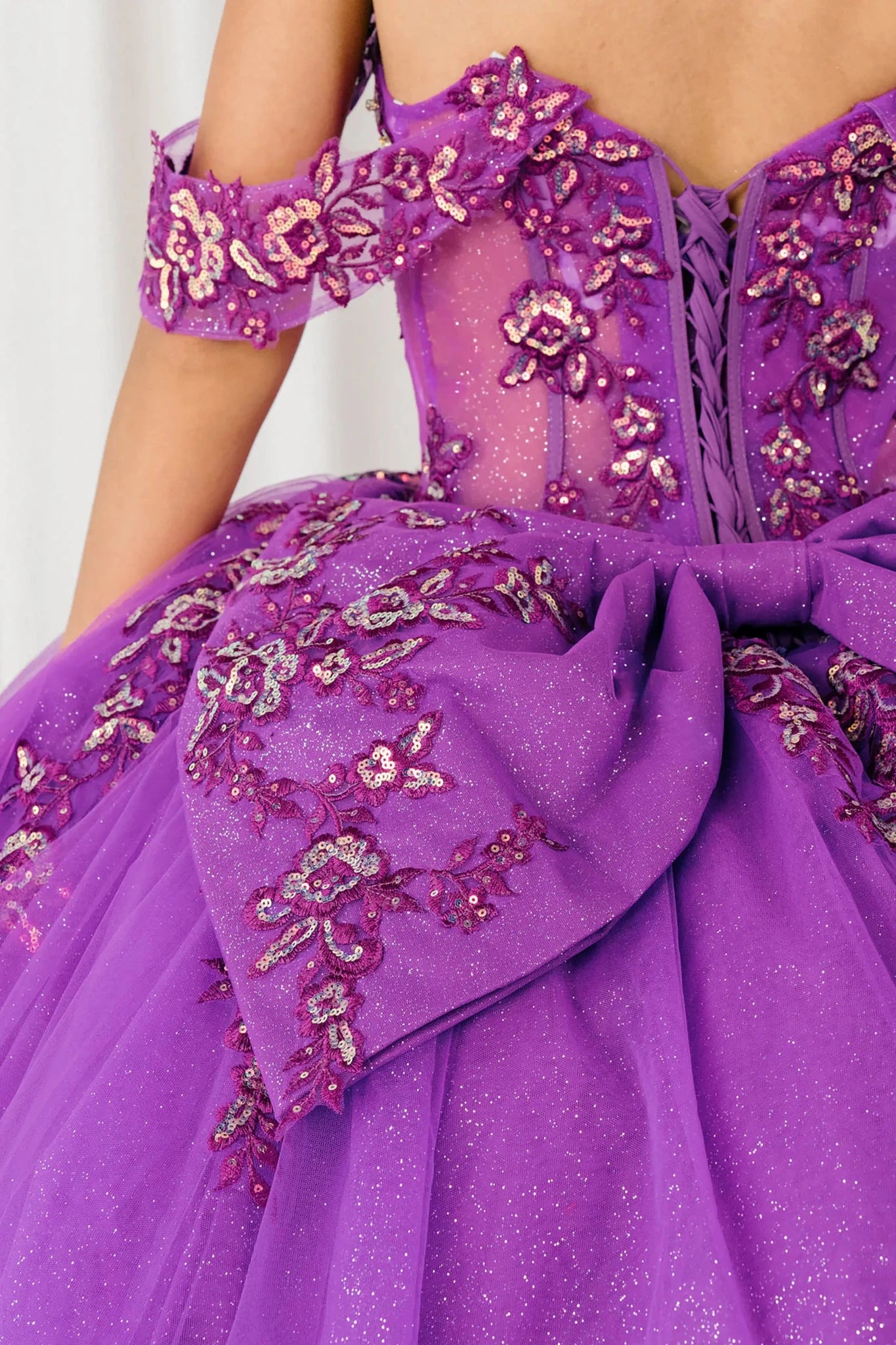 Back half close-up view of Magenta Off shoulder quinceañera ball gown with beaded corset, floral appliqués, layered tulle skirt, cathedral length train, and bow