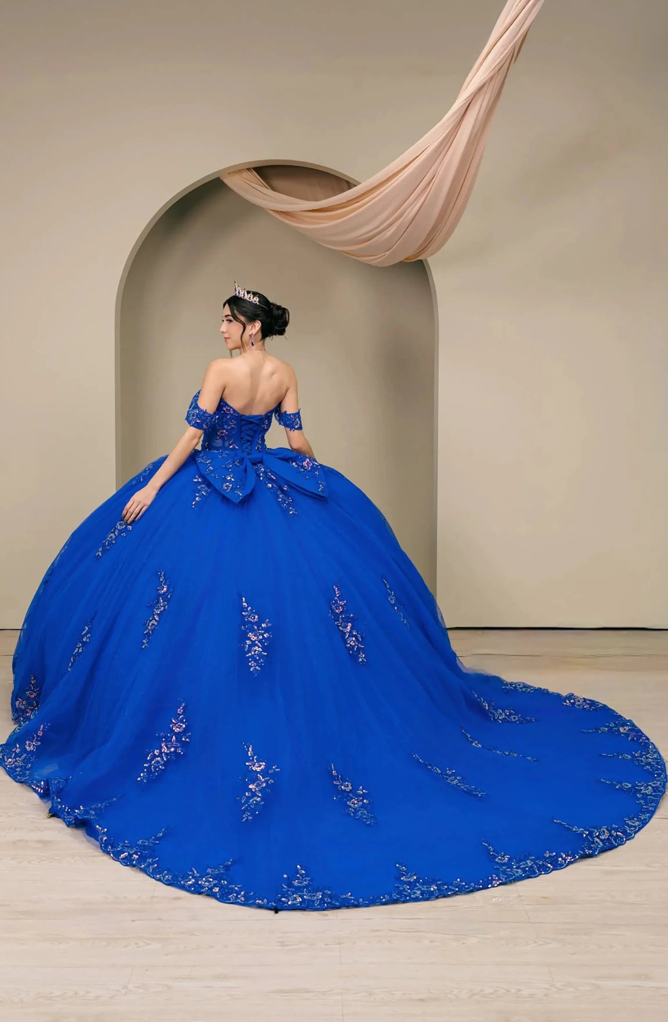 Back full-length view of Royal Off shoulder quinceañera ball gown with beaded corset, floral appliqués, layered tulle skirt, cathedral length train, and bow