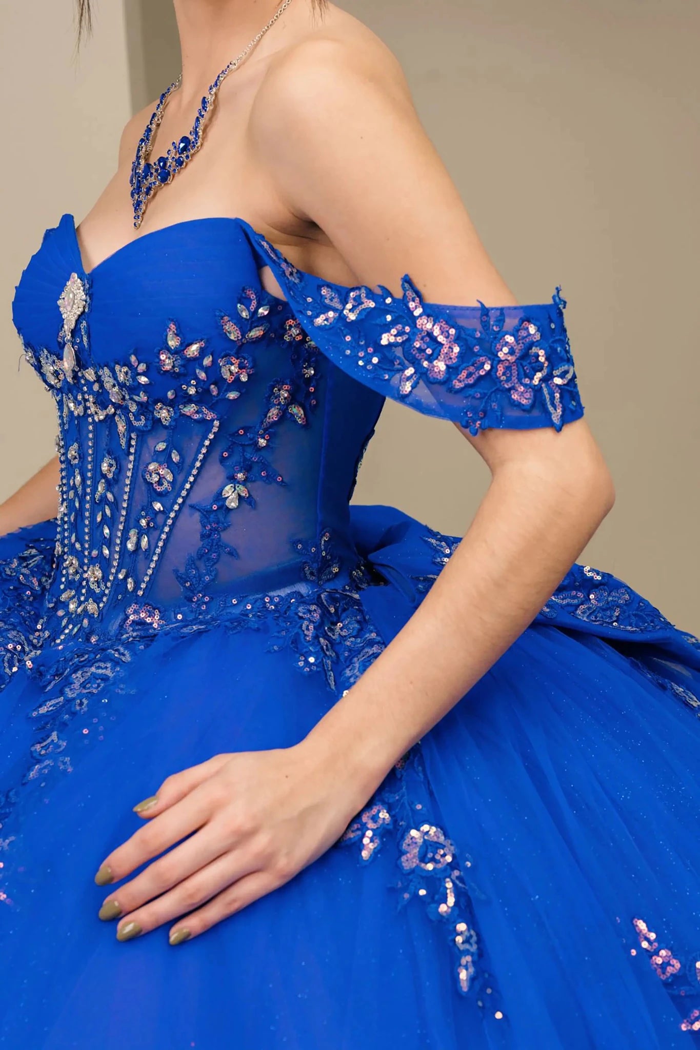 Side half close-up view of Royal Off shoulder quinceañera ball gown with beaded corset, floral appliqués, layered tulle skirt, cathedral length train, and bow
