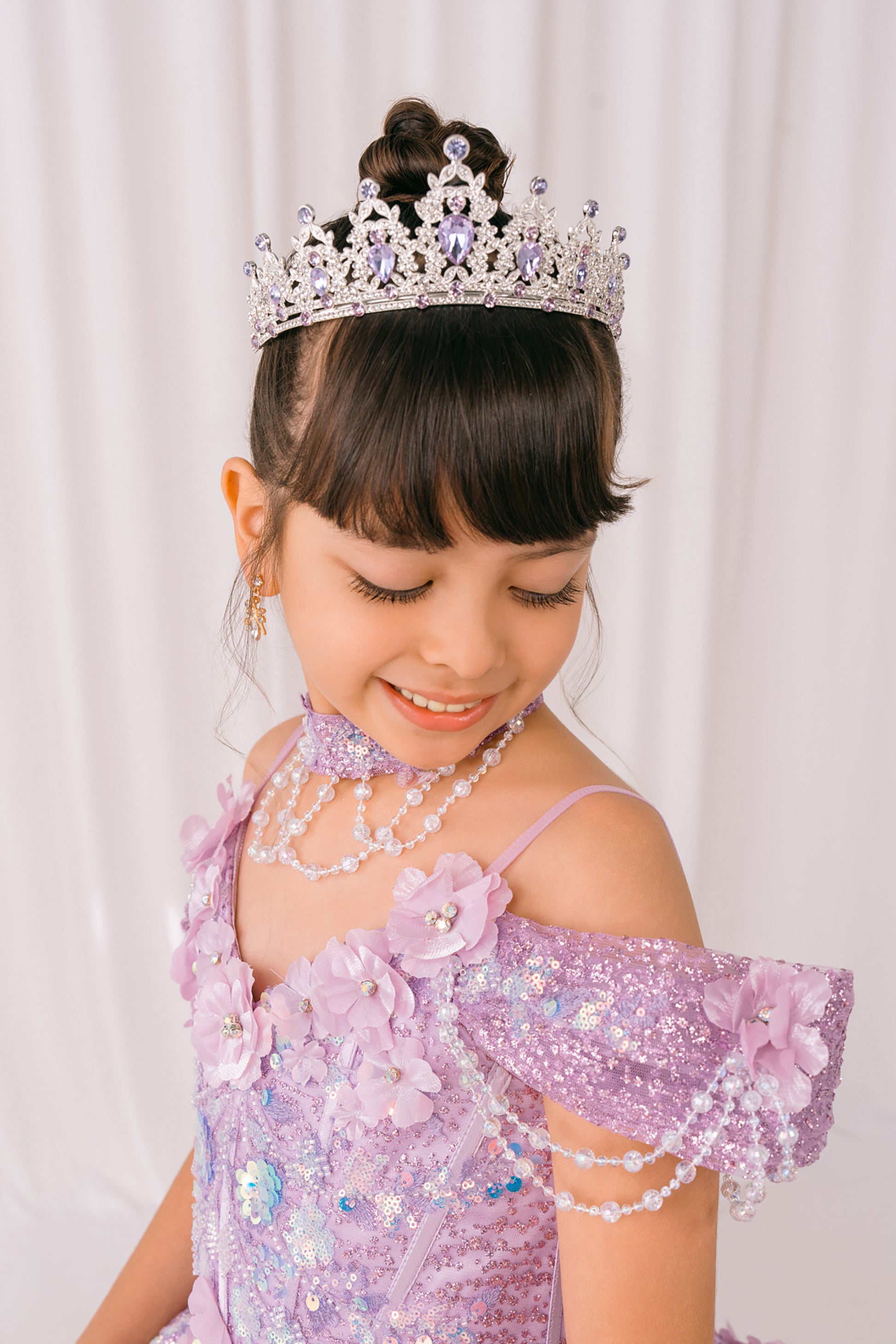 Young girl in a lilac mini quince dress with floral details and a tiara against a plain background