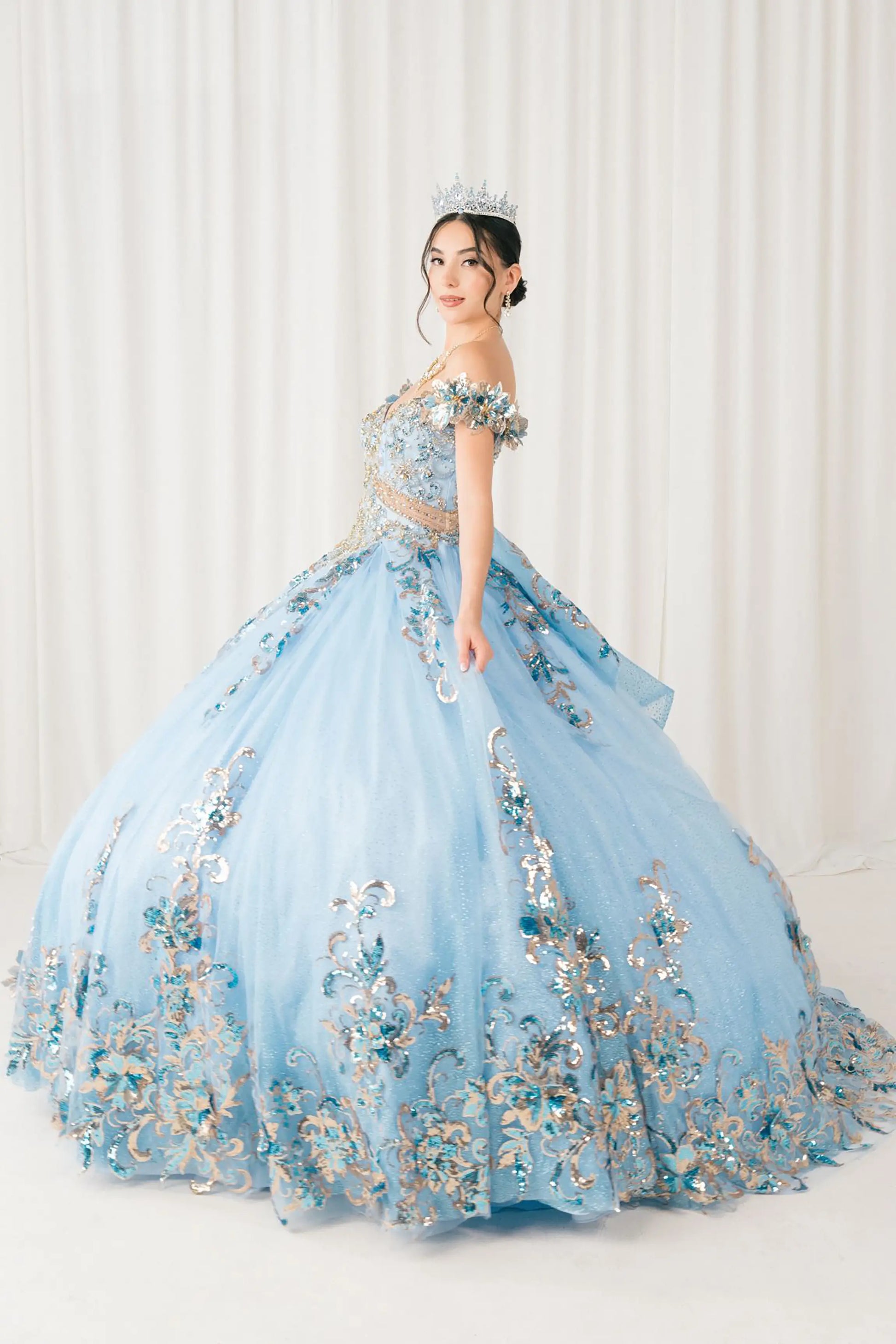 Side full-length view of Blue quinceañera ball gown with off shoulder bodice, gold embroidery, floral appliqués, voluminous tulle skirt, corset back, and oversized bow