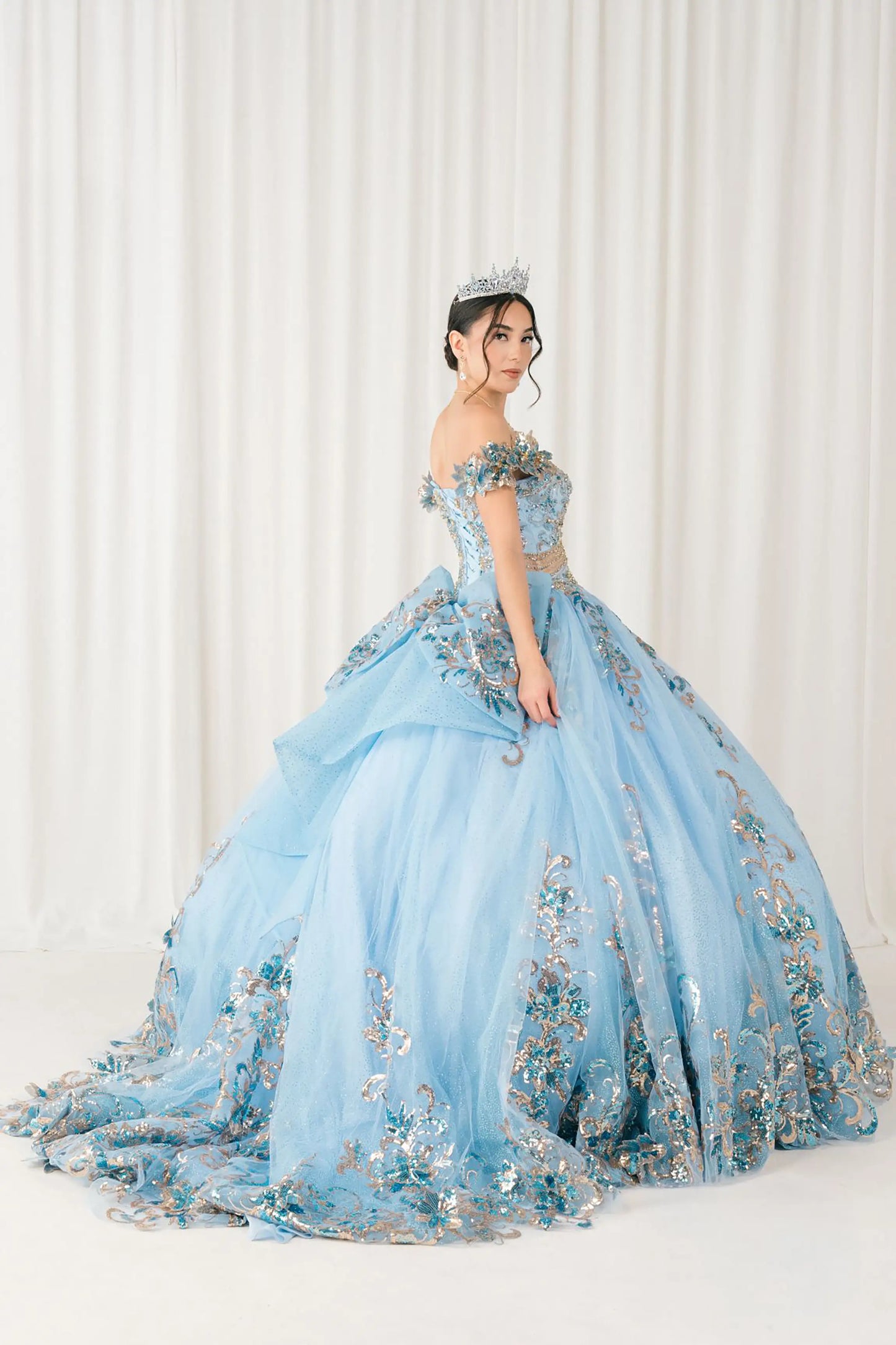 Side full-length view of Blue quinceañera ball gown with off shoulder bodice, gold embroidery, floral appliqués, voluminous tulle skirt, corset back, and oversized bow