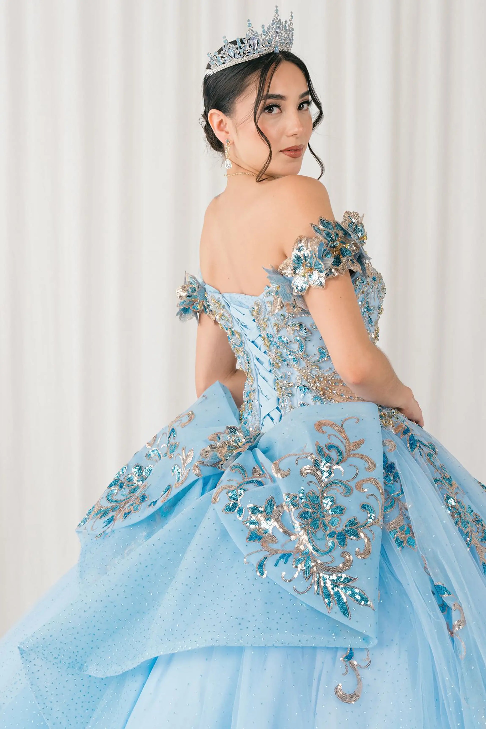 Side close-up view of Blue quinceañera ball gown with off shoulder bodice, gold embroidery, floral appliqués, voluminous tulle skirt, corset back, and oversized bow