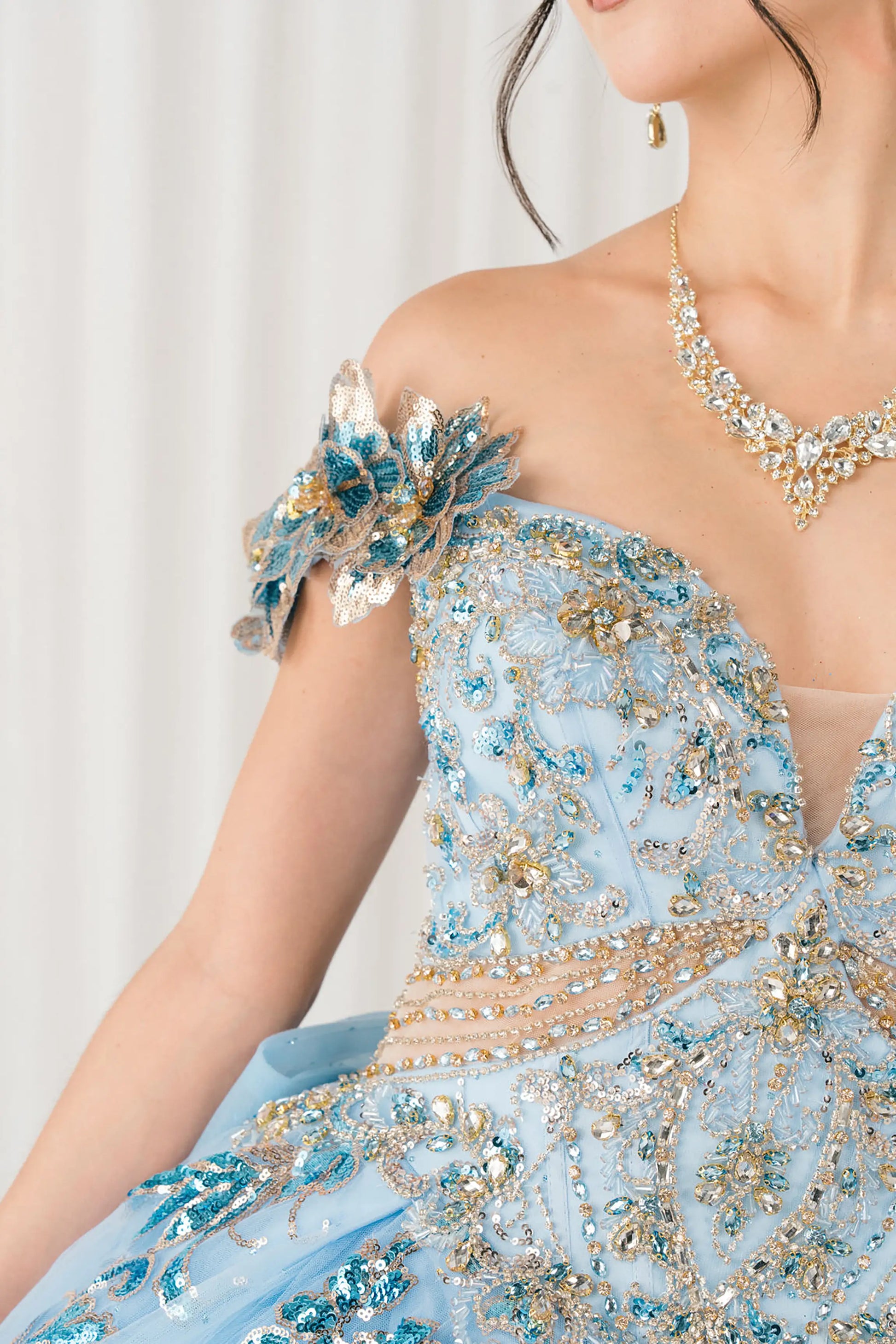 Side half close-up view of Blue quinceañera ball gown with off shoulder bodice, gold embroidery, floral appliqués, voluminous tulle skirt, corset back, and oversized bow