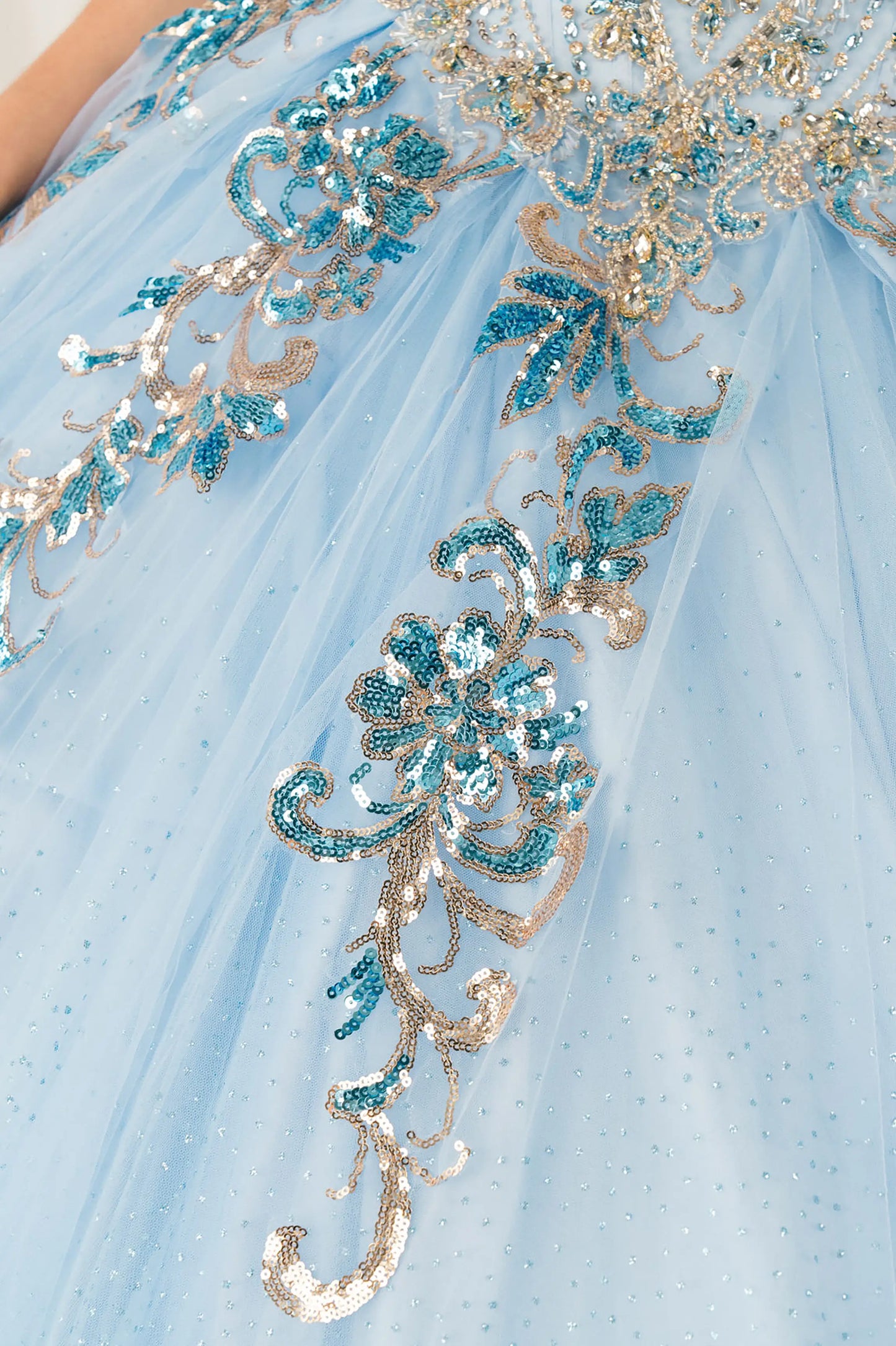 Close up view of Blue quinceañera ball gown with off shoulder bodice, gold embroidery, floral appliqués, voluminous tulle skirt, corset back, and oversized bow