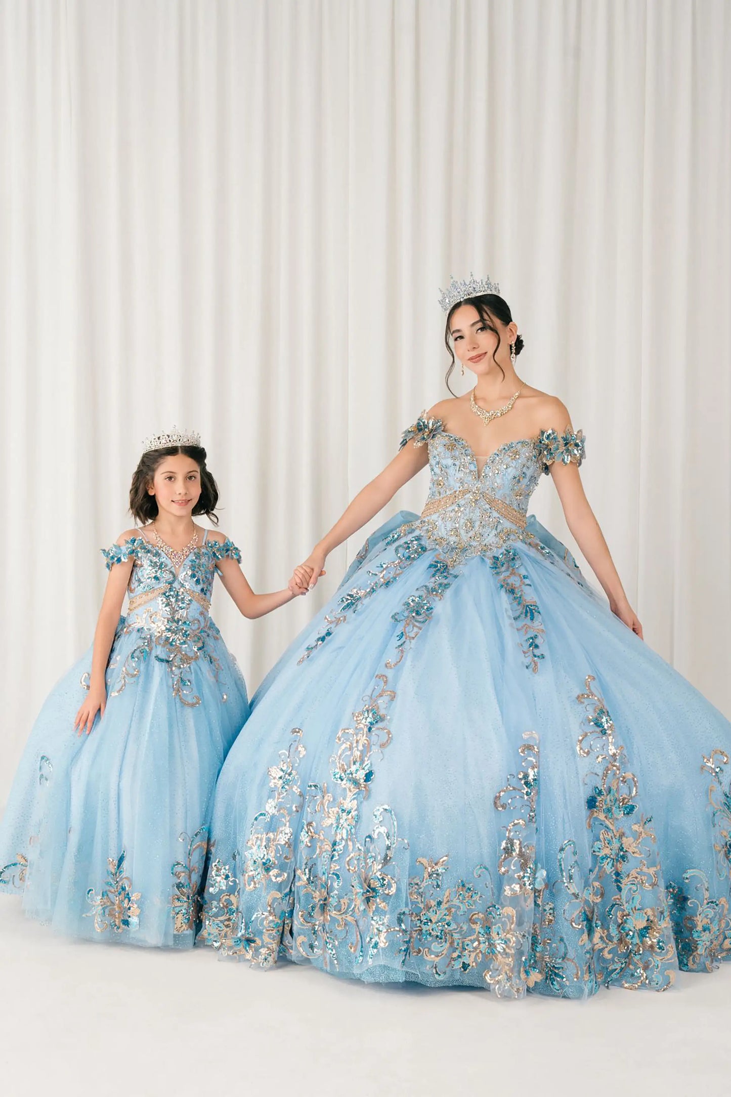 Blue quinceañera and mini quinceañera ball gown with off shoulder bodice, gold embroidery, floral appliqués, voluminous tulle skirt, corset back, and oversized bow