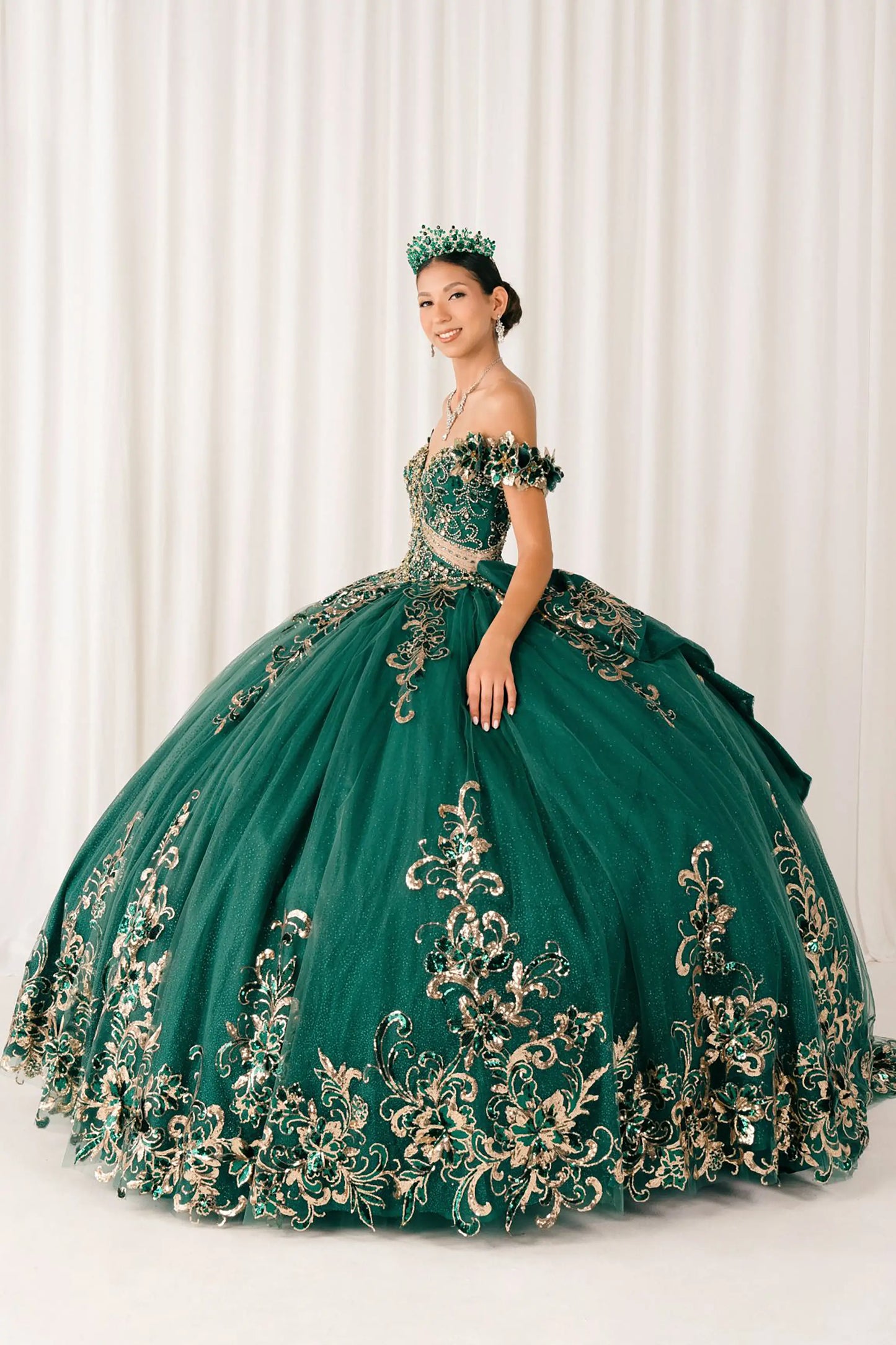 Side full-length view of Hunter Green quinceañera ball gown with off shoulder bodice, gold embroidery, floral appliqués, voluminous tulle skirt, corset back, and oversized bow