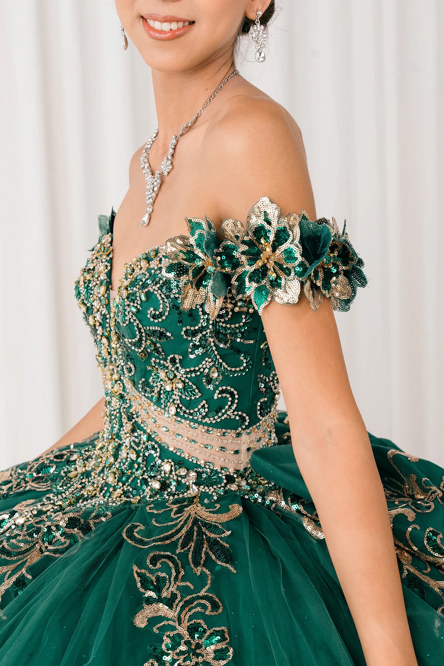 Side close-up Blue Hunter Green quinceañera ball gown with off shoulder bodice, gold embroidery, floral appliqués, voluminous tulle skirt, corset back, and oversized bow