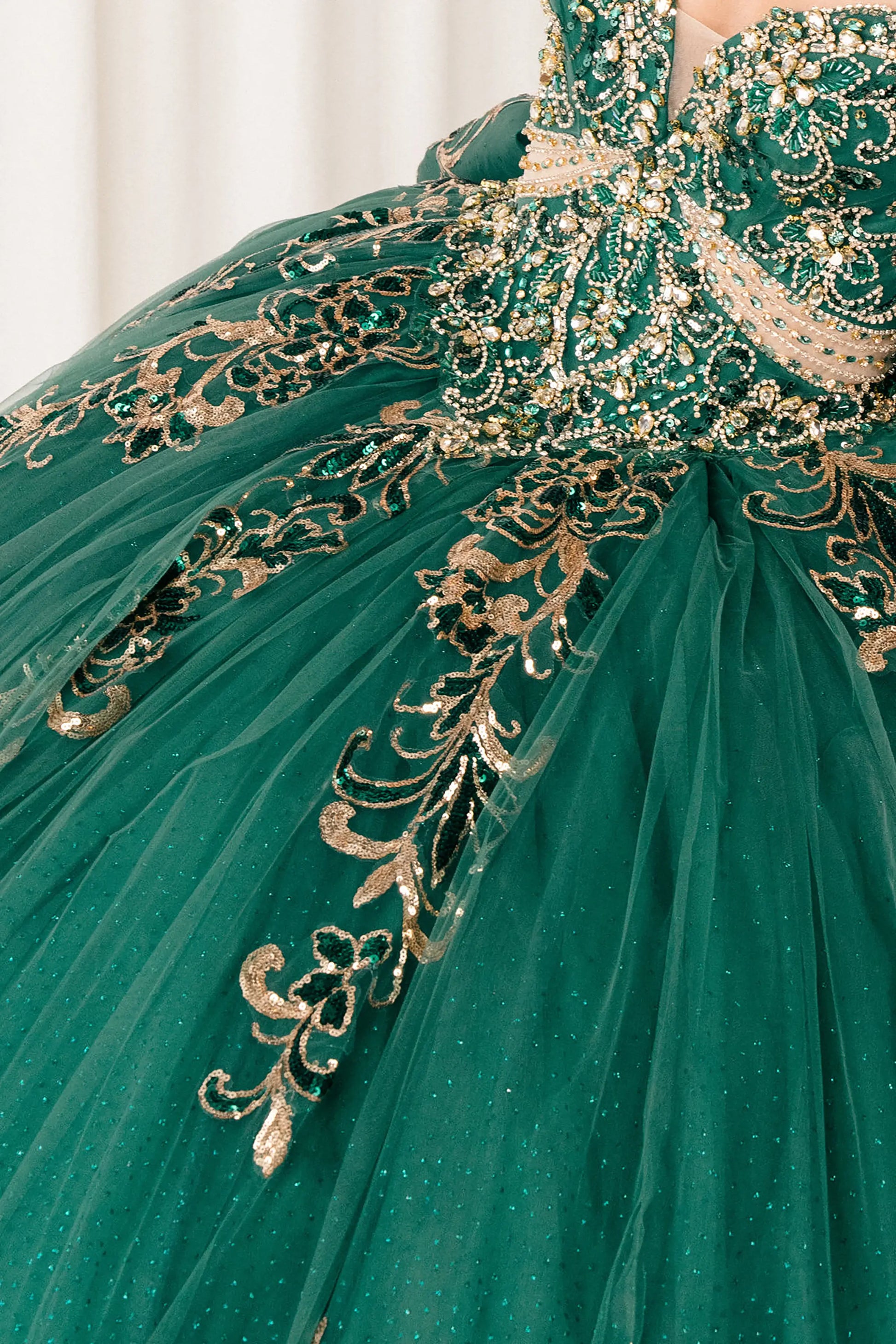 Close up Hunter Green quinceañera ball gown with off shoulder bodice, gold embroidery, floral appliqués, voluminous tulle skirt, corset back, and oversized bow
