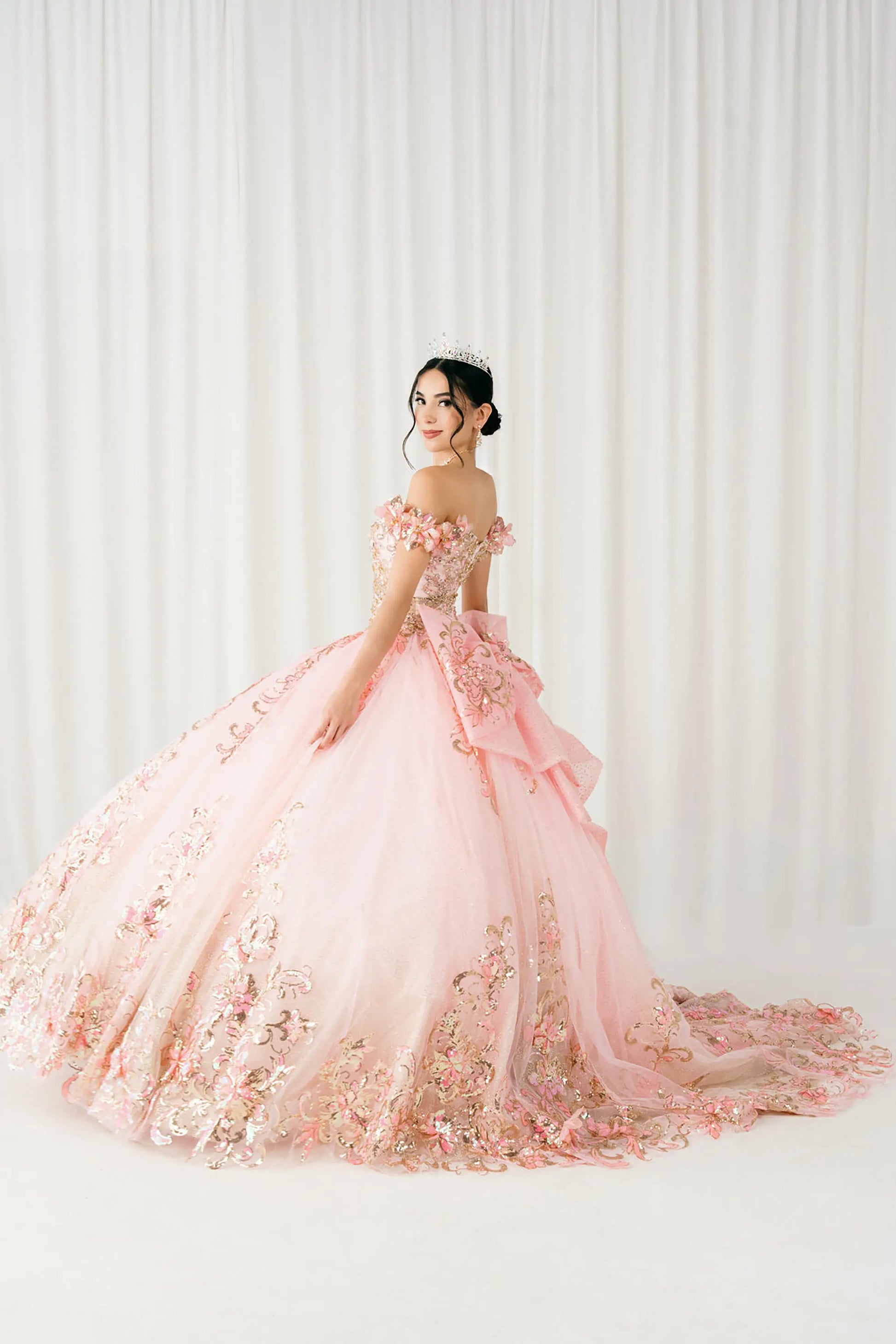 Side full-length view of Pink quinceañera ball gown with off shoulder bodice, gold embroidery, floral appliqués, voluminous tulle skirt, corset back, and oversized bow