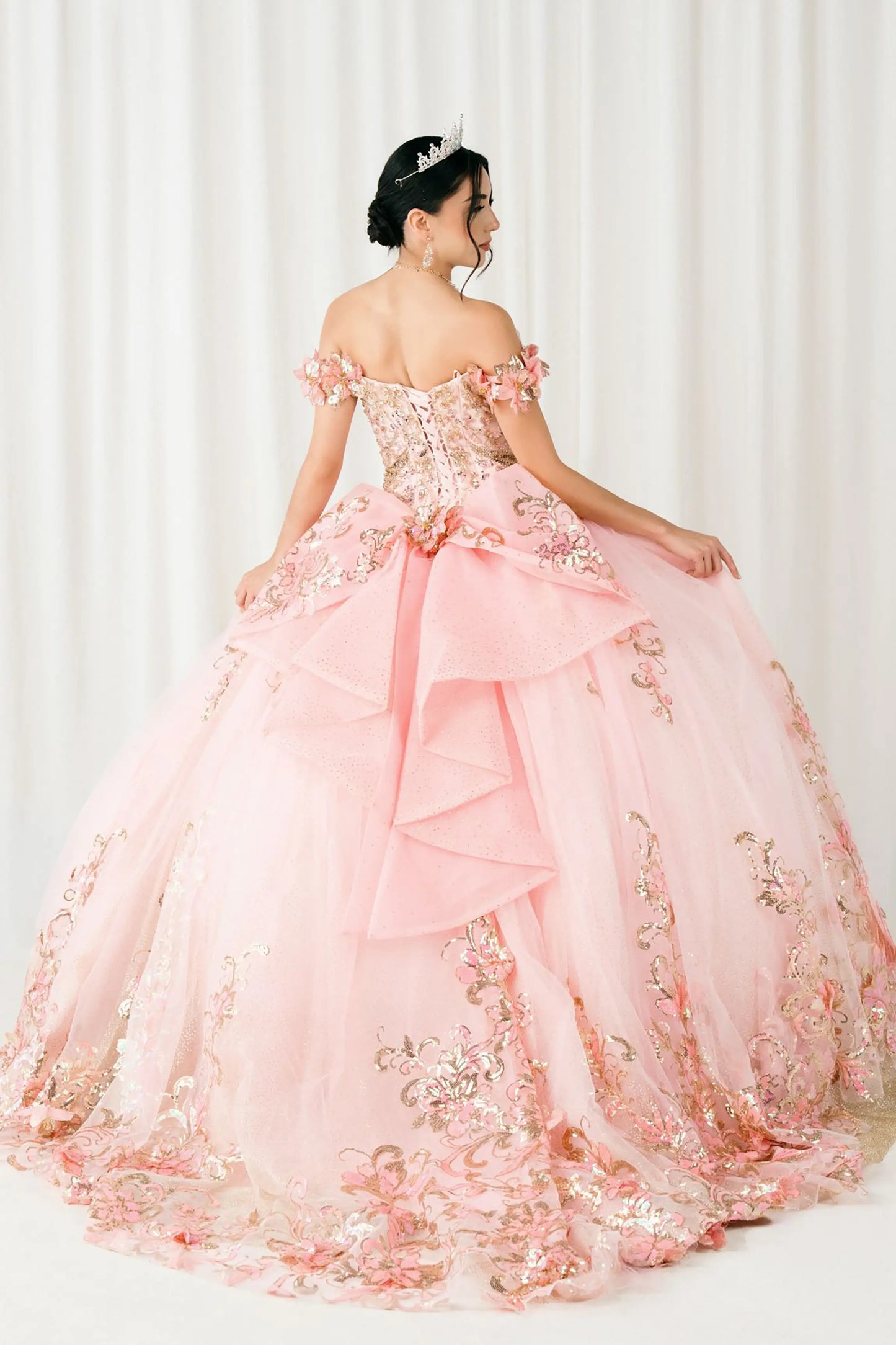 Back full-length view of Pink quinceañera ball gown with off shoulder bodice, gold embroidery, floral appliqués, voluminous tulle skirt, corset back, and oversized bow