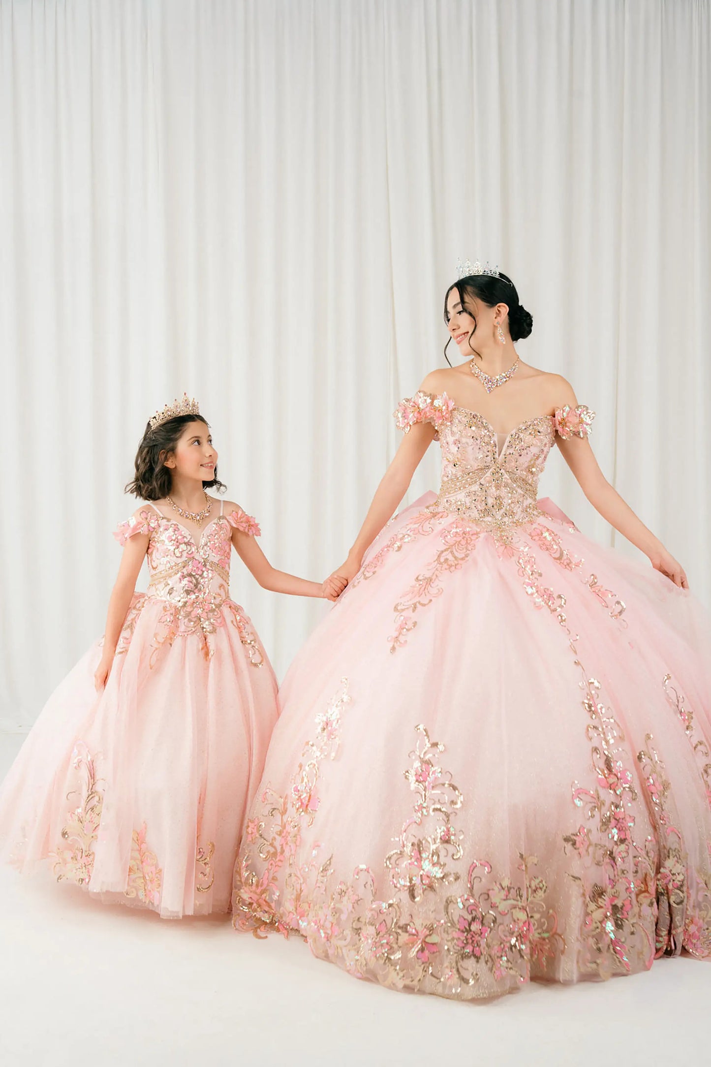 Pink quinceañera and mini quinceañera ball gown with off shoulder bodice, gold embroidery, floral appliqués, voluminous tulle skirt, corset back, and oversized bow