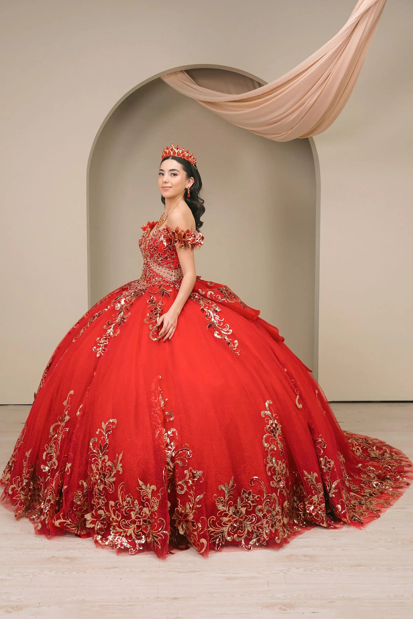 Side full-length view of Red quinceañera ball gown with off shoulder bodice, gold embroidery, floral appliqués, voluminous tulle skirt, corset back, and oversized bow