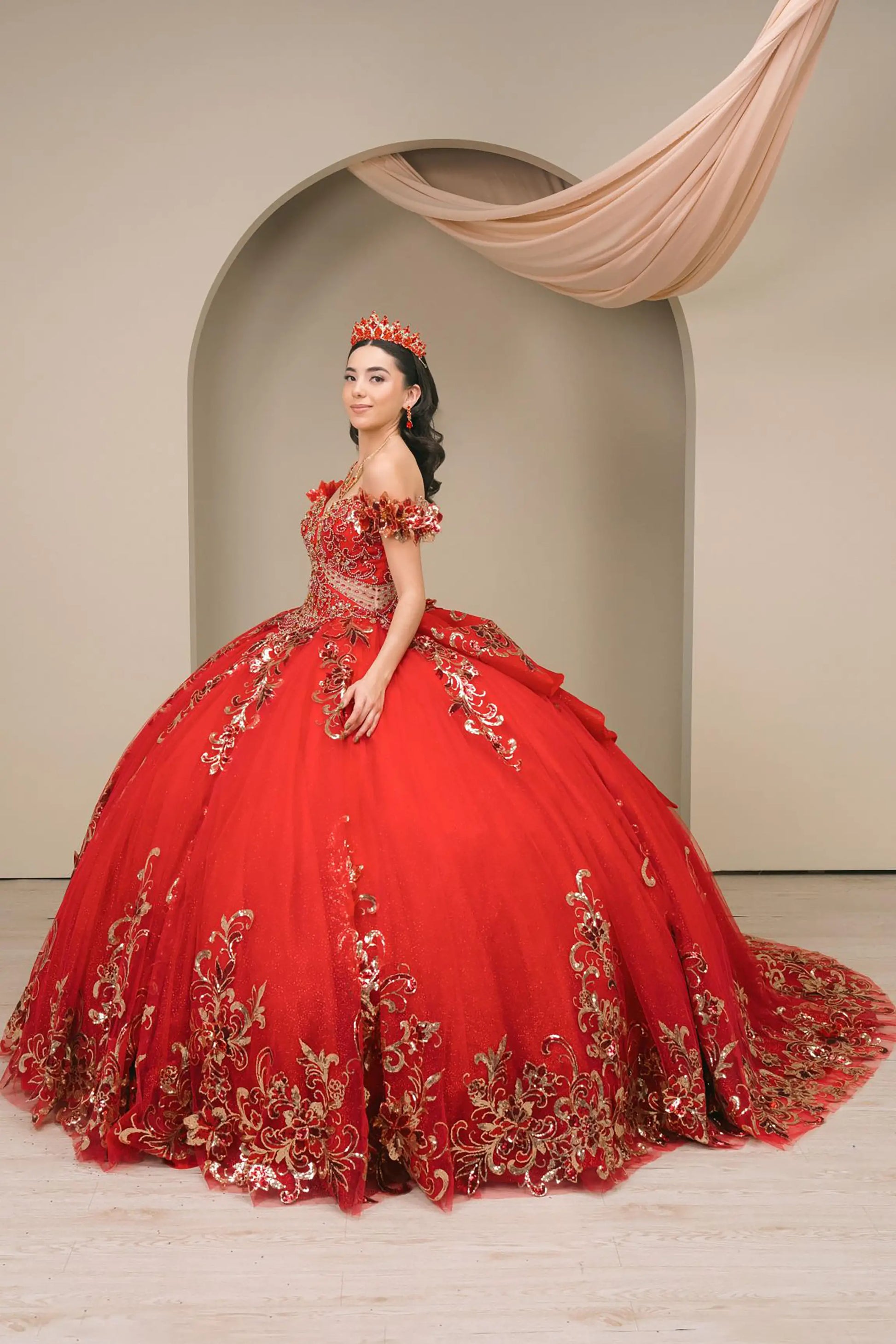 Side full-length view of Red quinceañera ball gown with off shoulder bodice, gold embroidery, floral appliqués, voluminous tulle skirt, corset back, and oversized bow