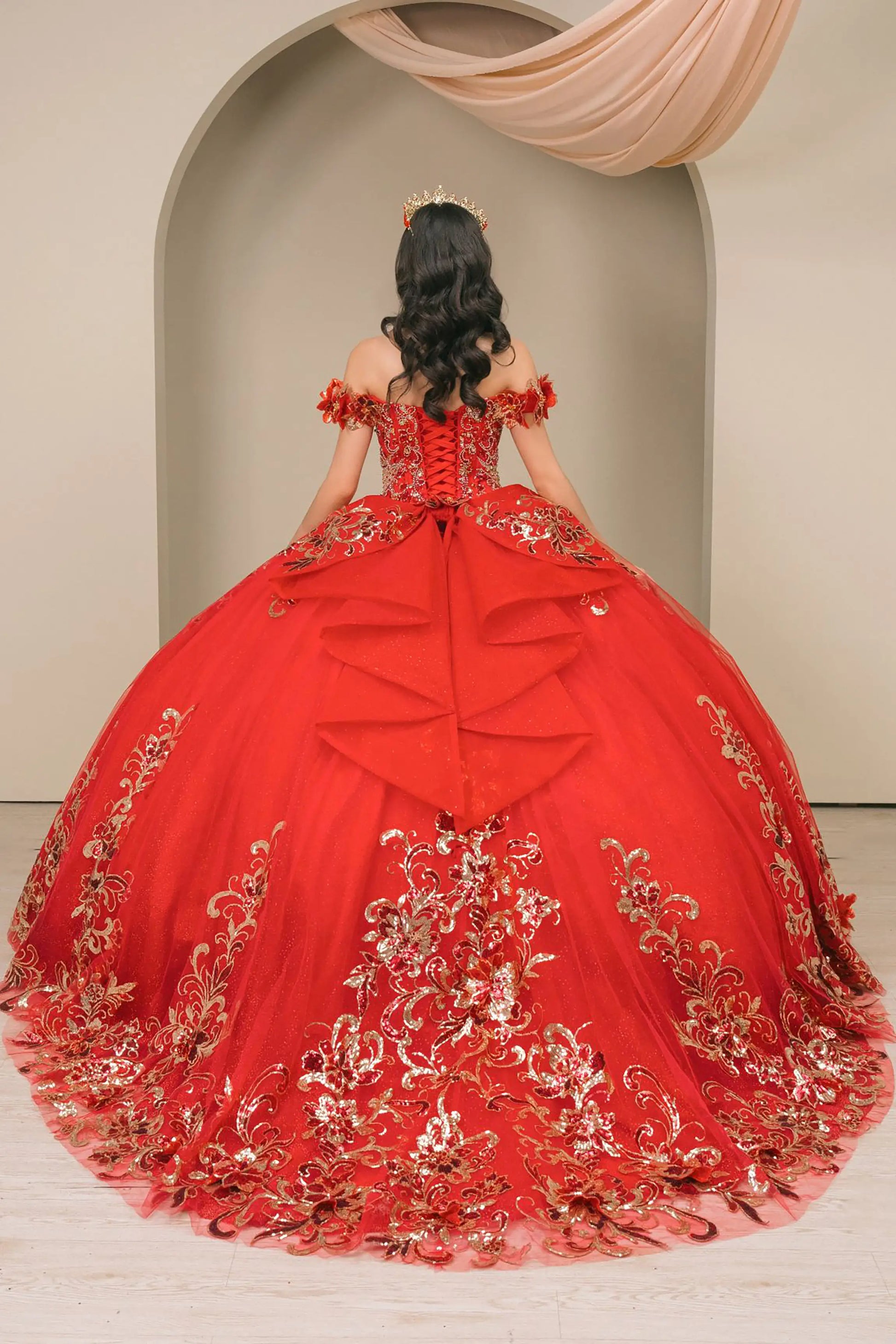 Back full-length view of Red quinceañera ball gown with off shoulder bodice, gold embroidery, floral appliqués, voluminous tulle skirt, corset back, and oversized bow