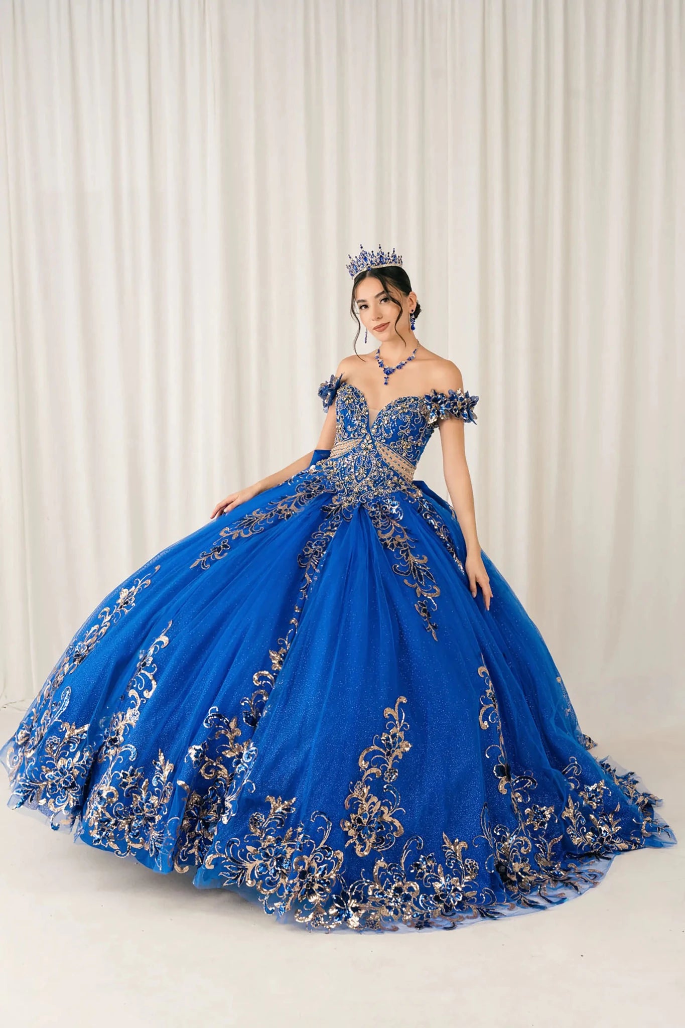 Front full-length view of Royal quinceañera ball gown with off shoulder bodice, gold embroidery, floral appliqués, voluminous tulle skirt, corset back, and oversized bow