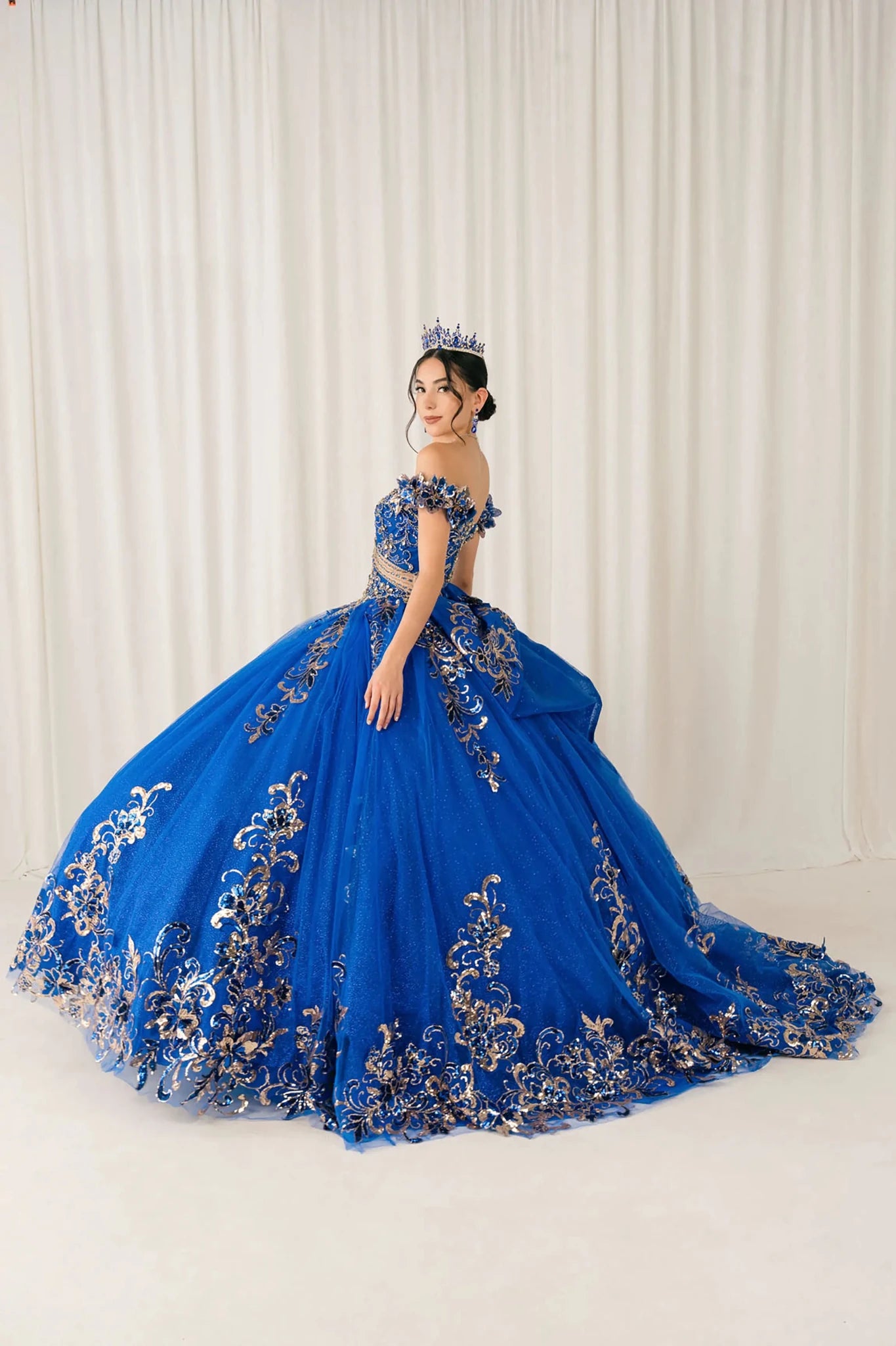 Side full-length view of Royal quinceañera ball gown with off shoulder bodice, gold embroidery, floral appliqués, voluminous tulle skirt, corset back, and oversized bow