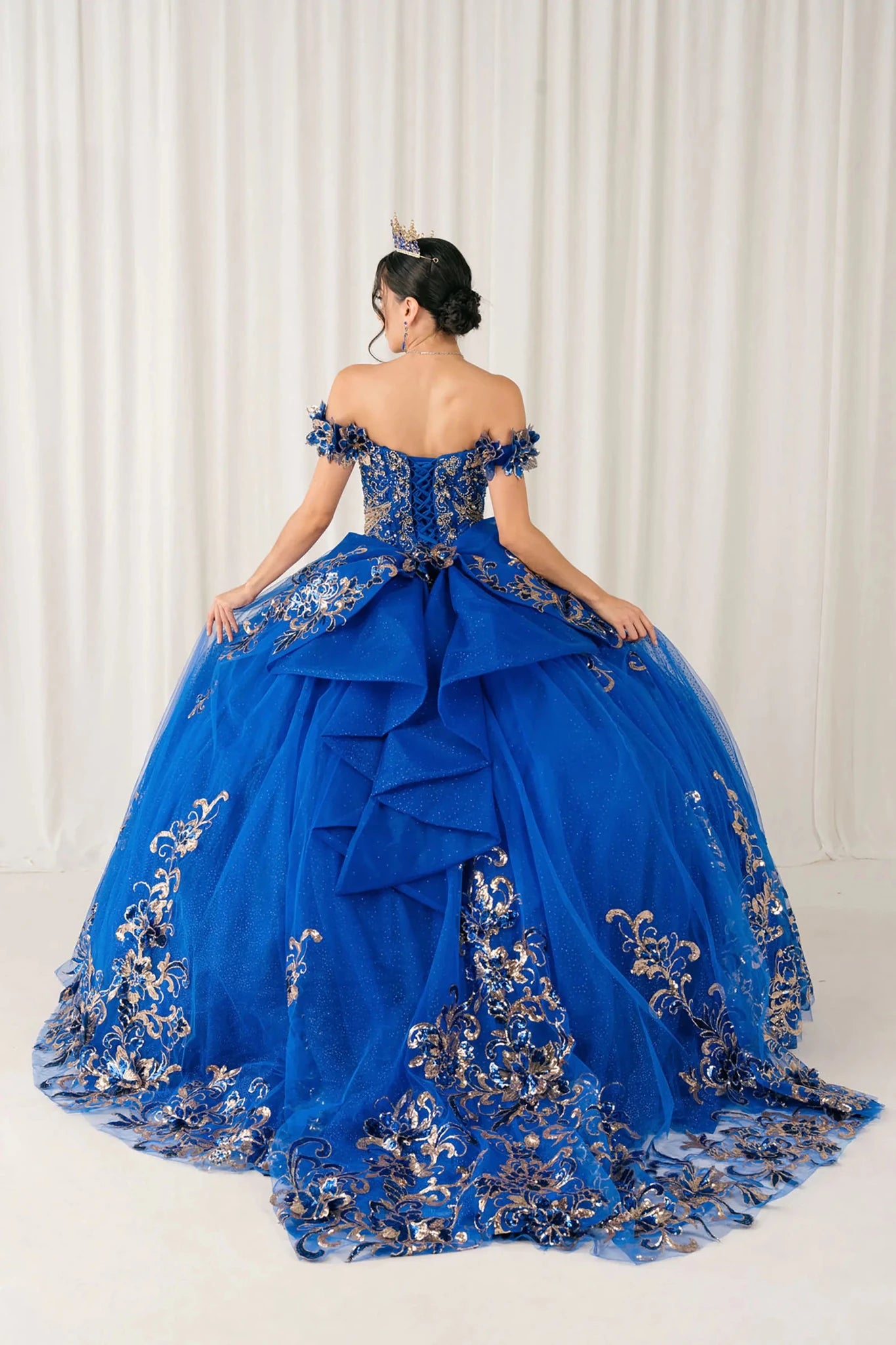 Back full-length view of Royal quinceañera ball gown with off shoulder bodice, gold embroidery, floral appliqués, voluminous tulle skirt, corset back, and oversized bow