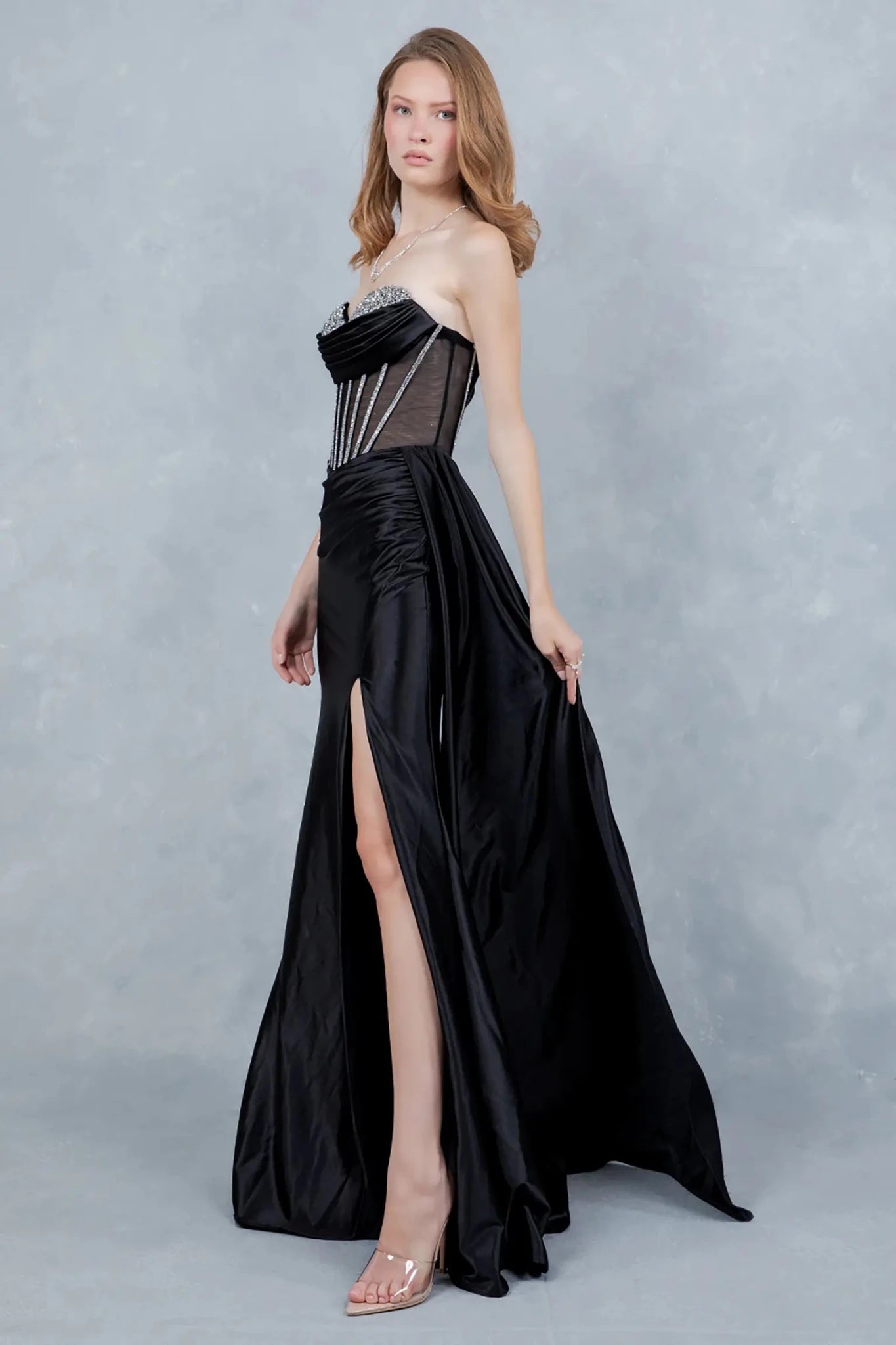 A side profile view of a woman wearing a black satin prom dress, highlighting the ruched waist, fitted silhouette, and slit.