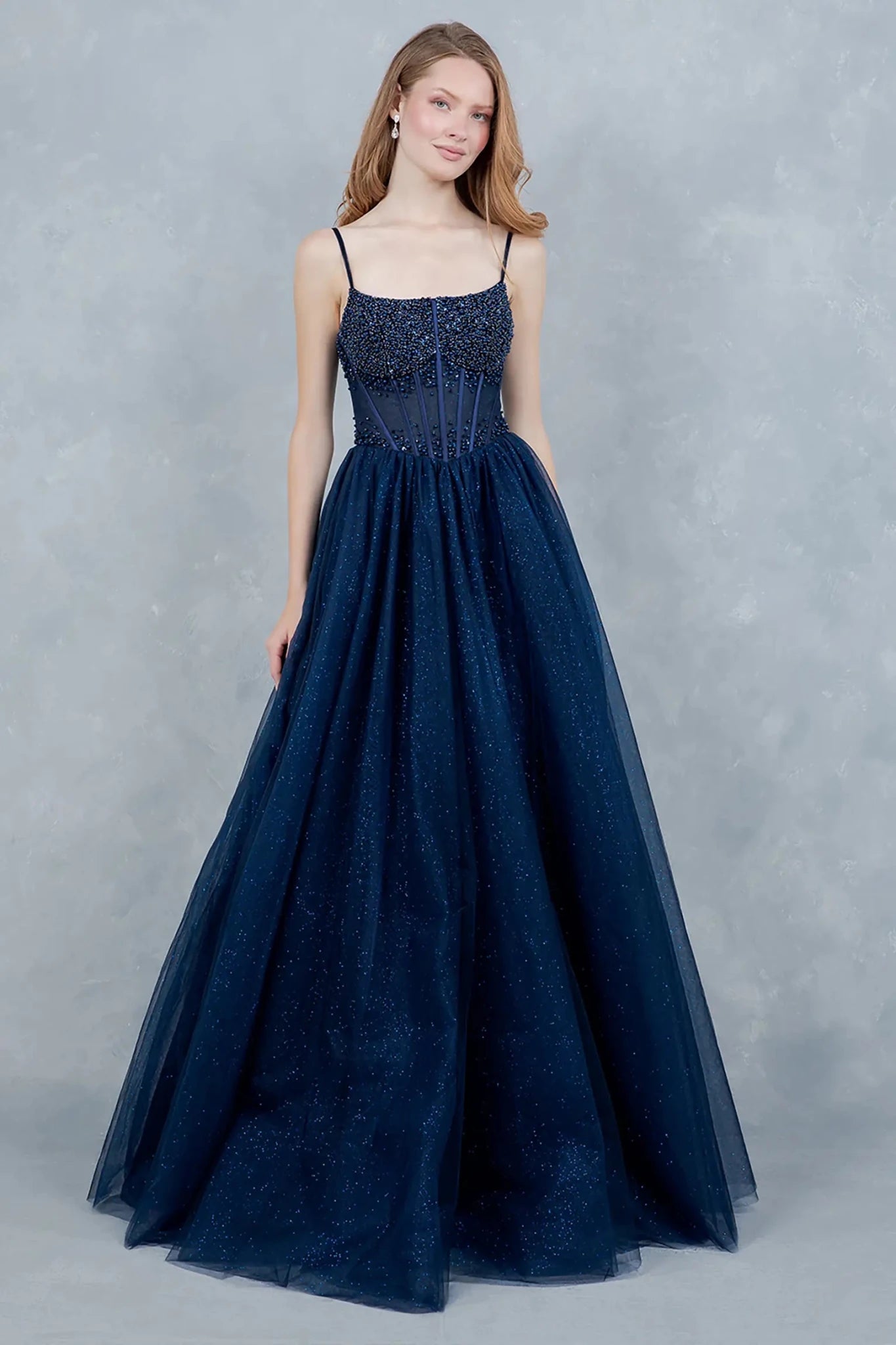 A full-length front view of a woman gracefully wearing a navy fairytale prom dress featuring a lace-up corset and glitter tulle skirt.
