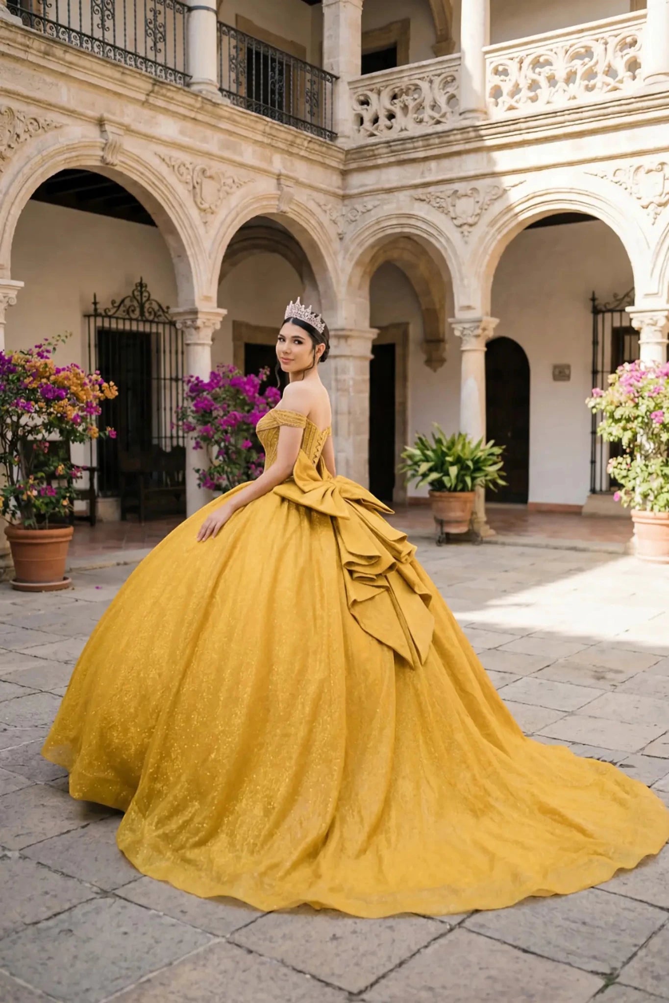 Woman wearing gold off-shoulder ball gown side-facing with beaded corset bodice, shimmering glitter tulle layers, and flowing silhouette