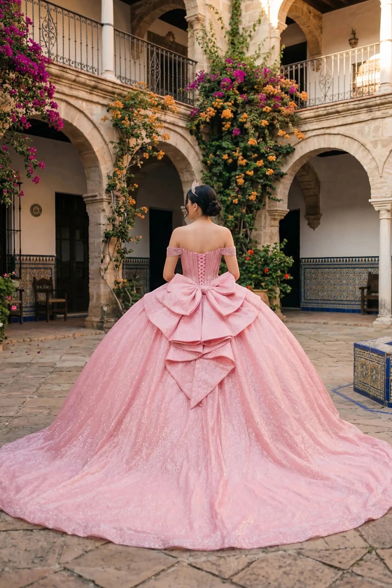 Woman back-facing in rose gold off-shoulder ball gown with lace-up corset bodice, intricate beadwork, glitter tulle, and cascading full skirt