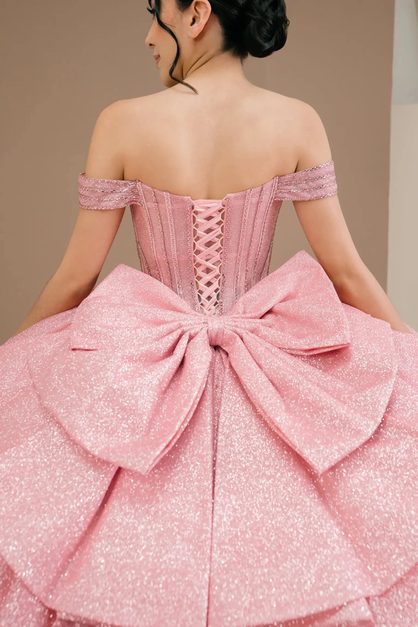 Woman Close up back-facing in rose gold off-shoulder ball gown with lace-up corset bodice, intricate beadwork, glitter tulle, and cascading full skirt
