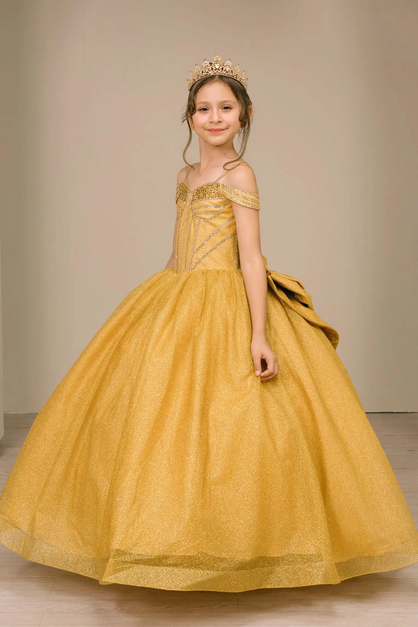 Kid standing back-facing in gold ball gown with lace-up closure, oversized tiered bow centerpiece, and cascading layered skirt

