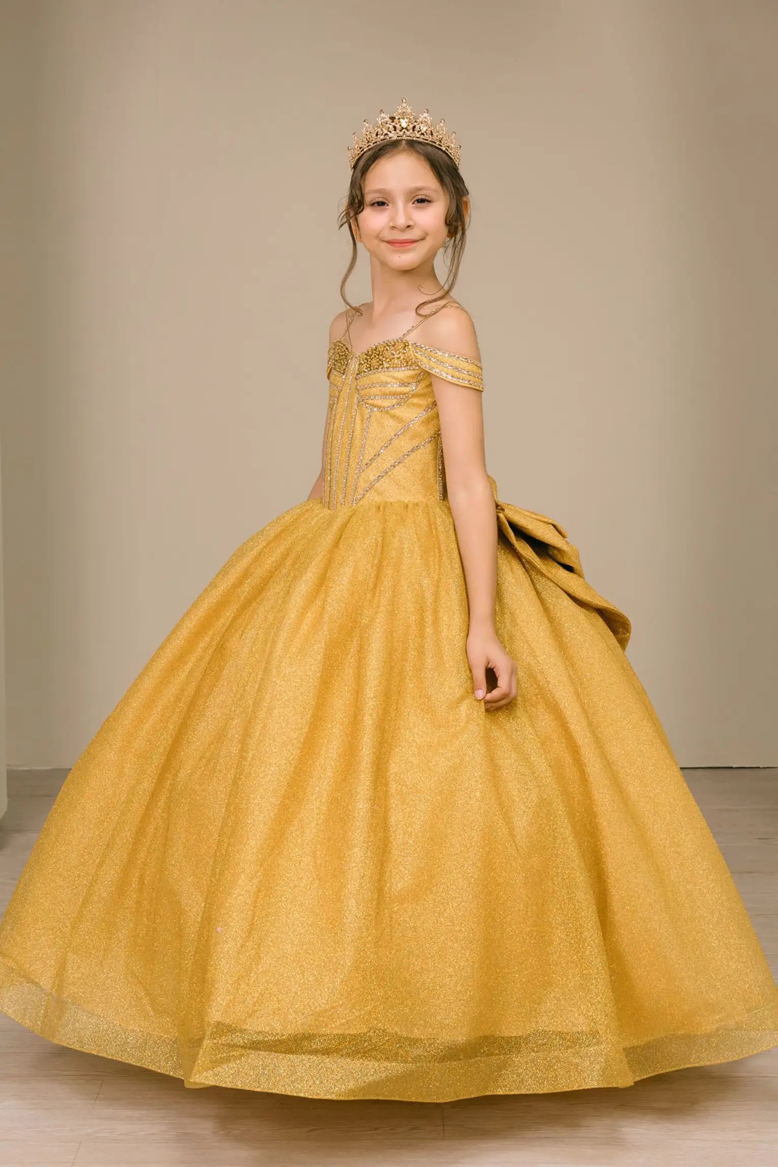 Kid standing back-facing in gold ball gown with lace-up closure, oversized tiered bow centerpiece, and cascading layered skirt


