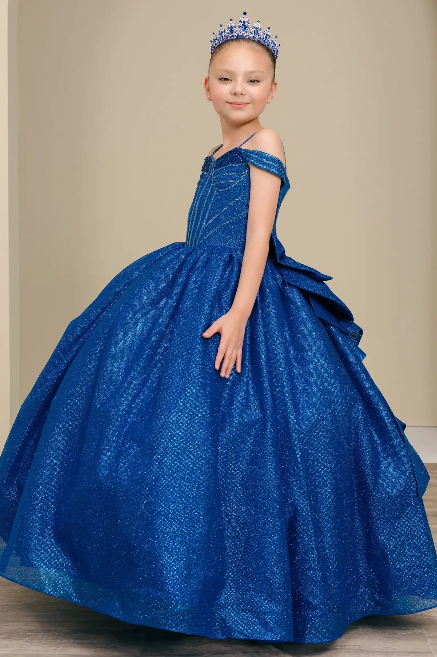 Kid standing side-facing in royal blue ball gown with oversized tiered bow detail, shimmering bodice, and flowing layered skirt

