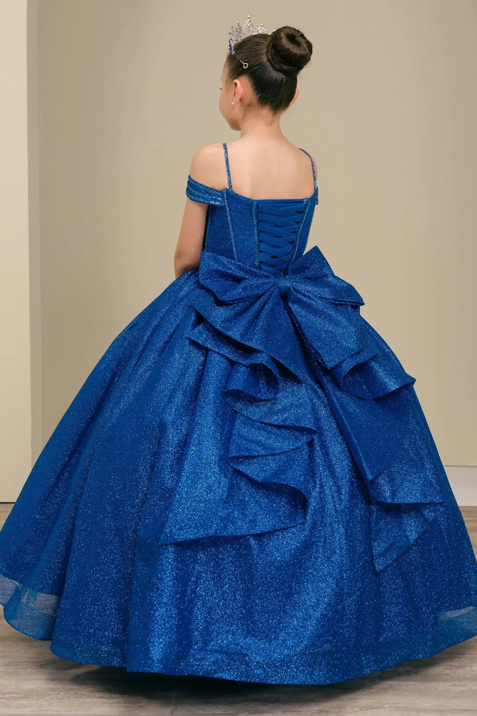 Kid standing three-quarter view in royal blue ball gown with oversized tiered bow, glittering bodice, and dramatic layered skirt

