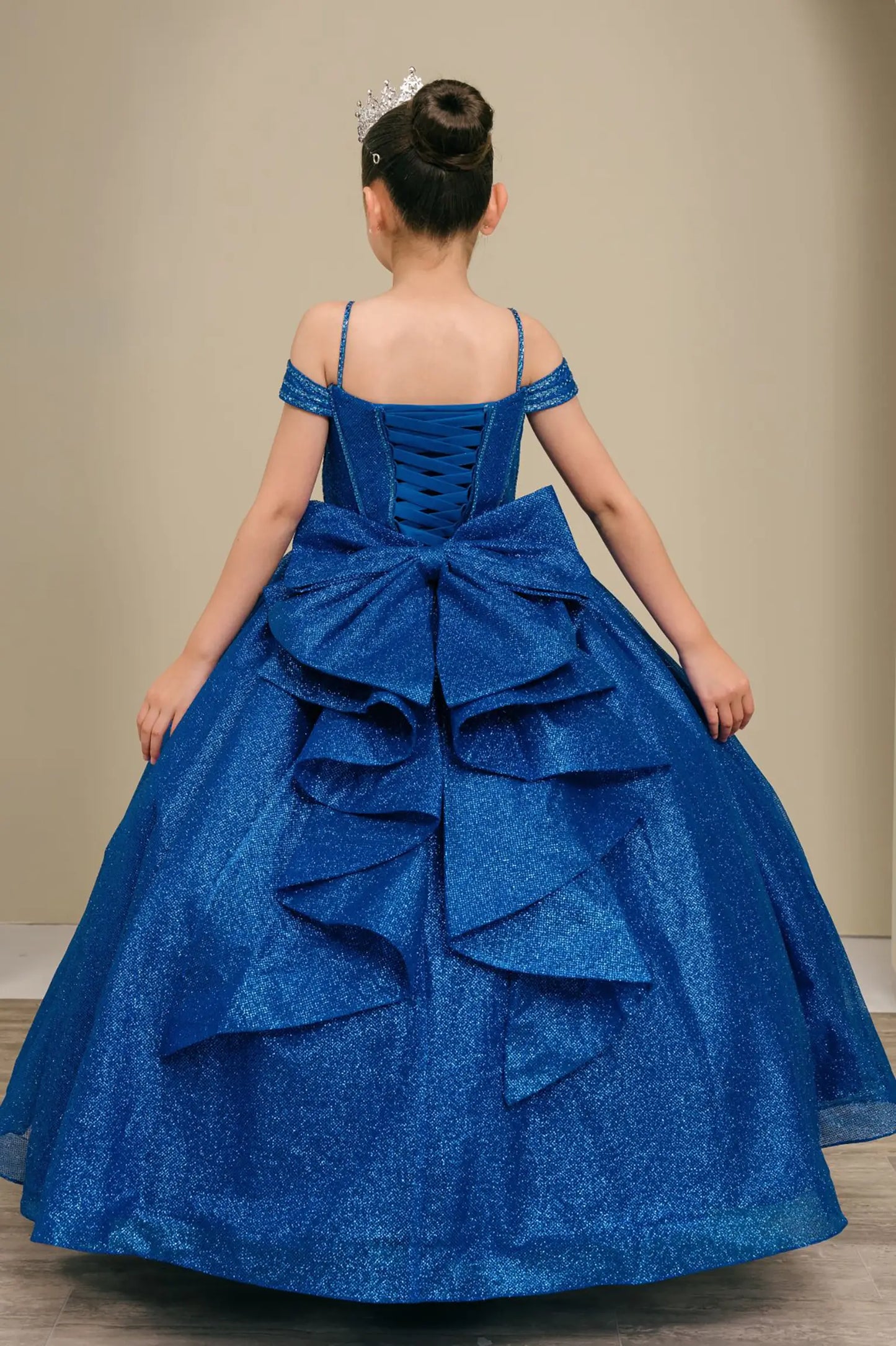 Kid standing side-facing in royal blue ball gown with oversized tiered bow detail, shimmering bodice, and flowing layered skirt

