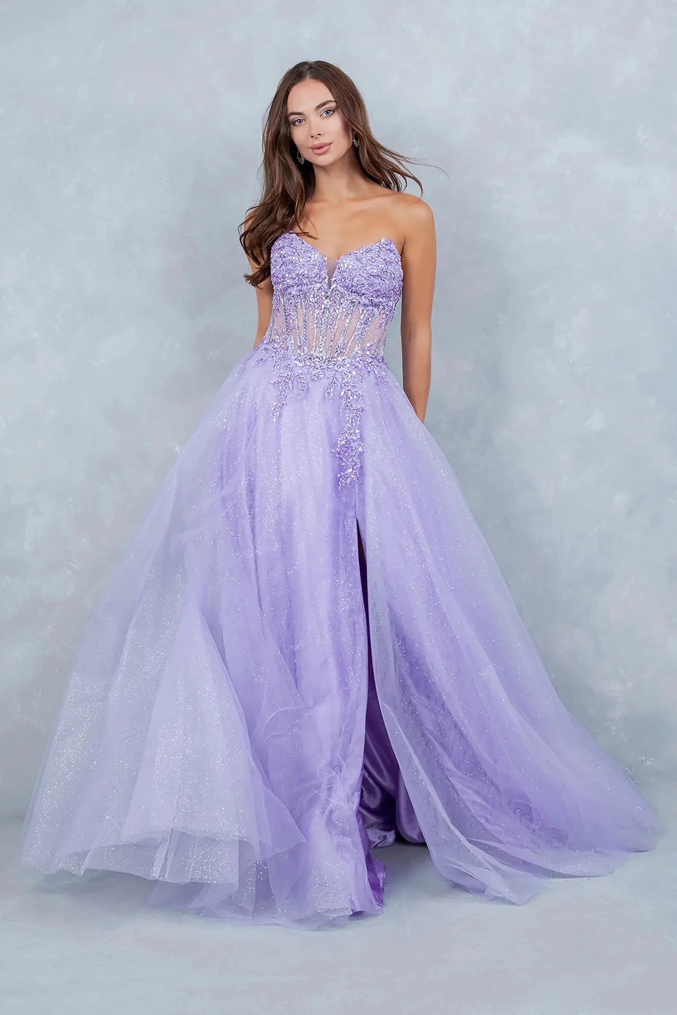 A full-length front view of a woman gracefully wearing a lilac fairytale prom dress featuring a lace-up corset and a layered glitter tulle skirt.