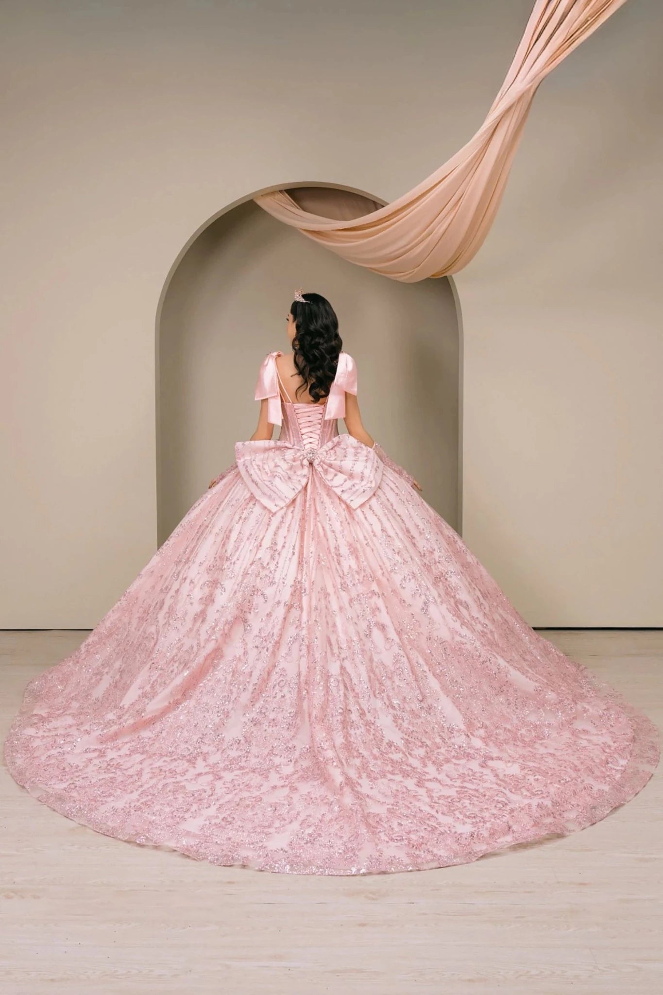 Woman back-facing in blush off-shoulder sweetheart quinceañera dress with lace-up beaded corset bodice, lace appliqué, oversized back bow, and cascading layered tulle skirt