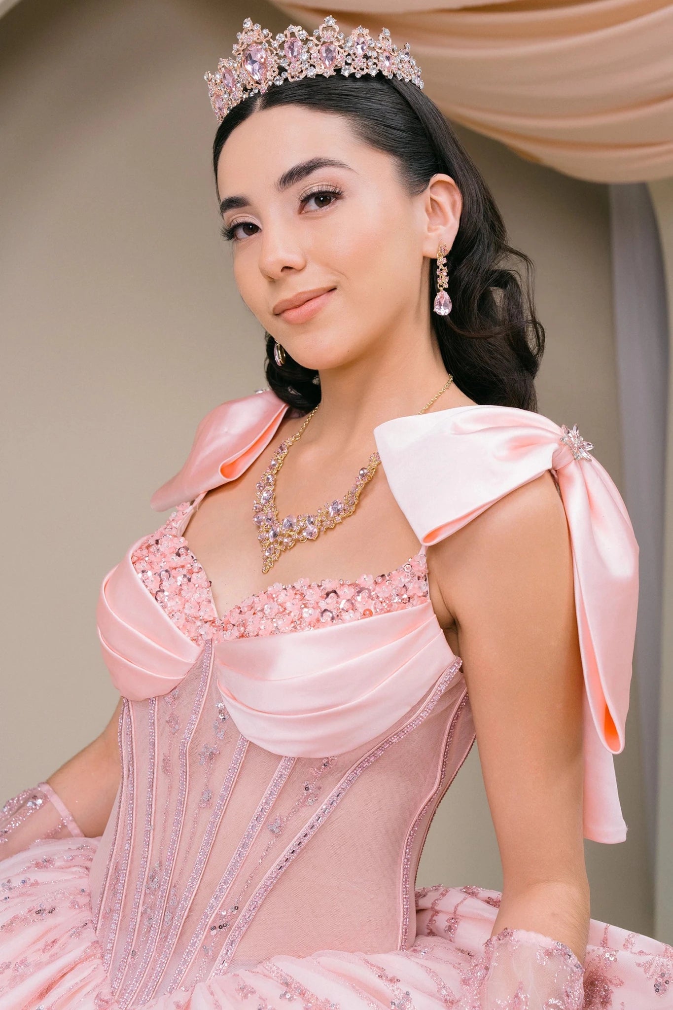 Woman wearing blush off-shoulder sweetheart quinceañera dress three-quarter view with ornate beaded corset bodice, lace appliqué, layered tulle skirt, and back bow accent