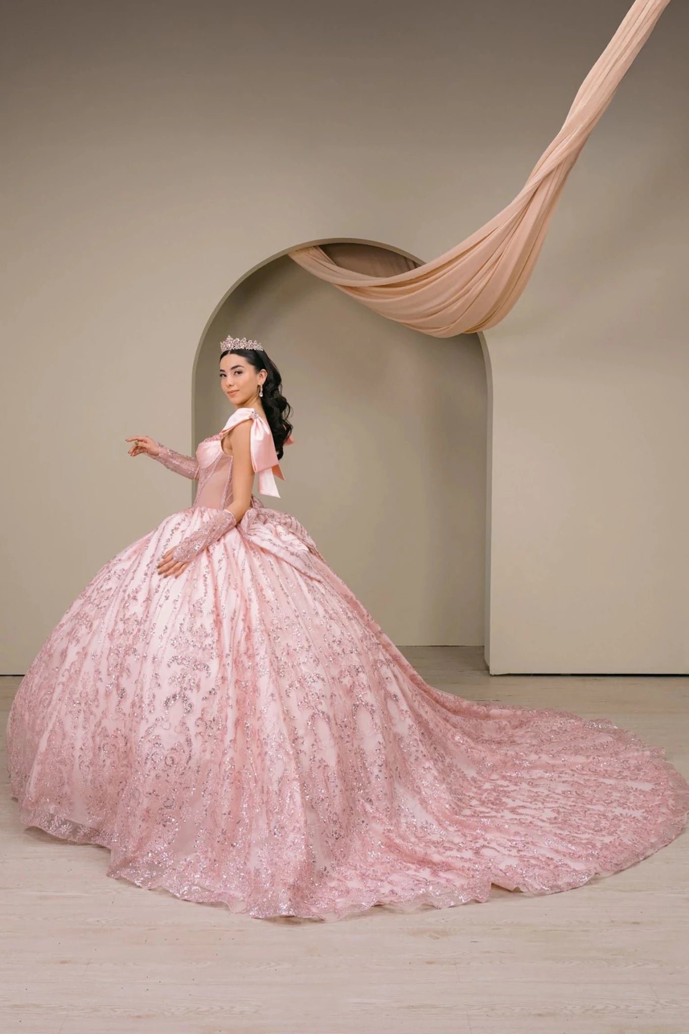 Woman wearing blush off-shoulder sweetheart quinceañera dress side-facing with beaded corset bodice, lace appliqué, layered tulle skirt, and back bow accent